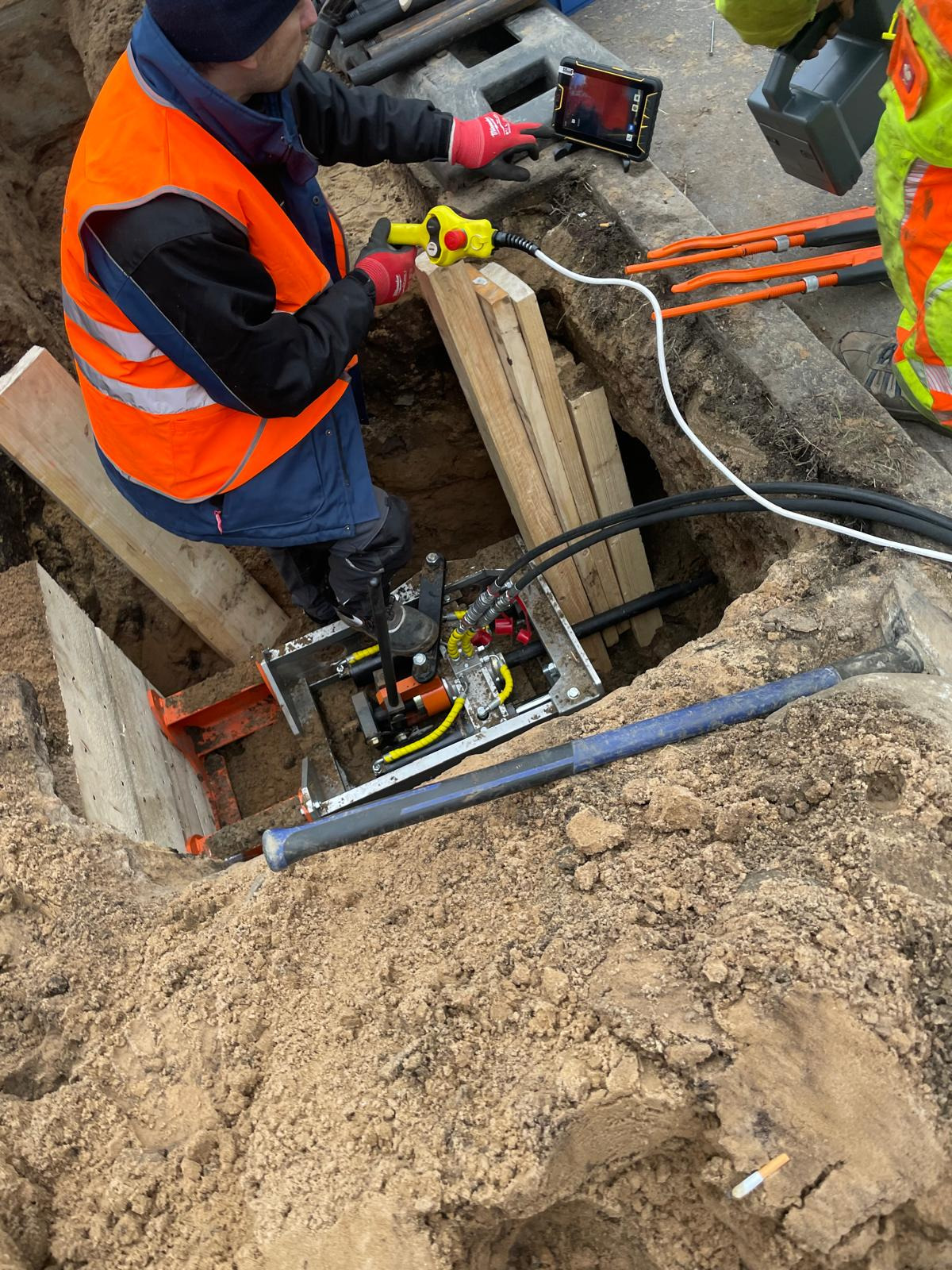 Pracownik w kamizelce odblaskowej obsługuje urządzenie do inspekcji rur w wykopie zabezpieczonym drewnianą konstrukcją, używając tabletu do podglądu obrazu z kamery inspekcyjnej.