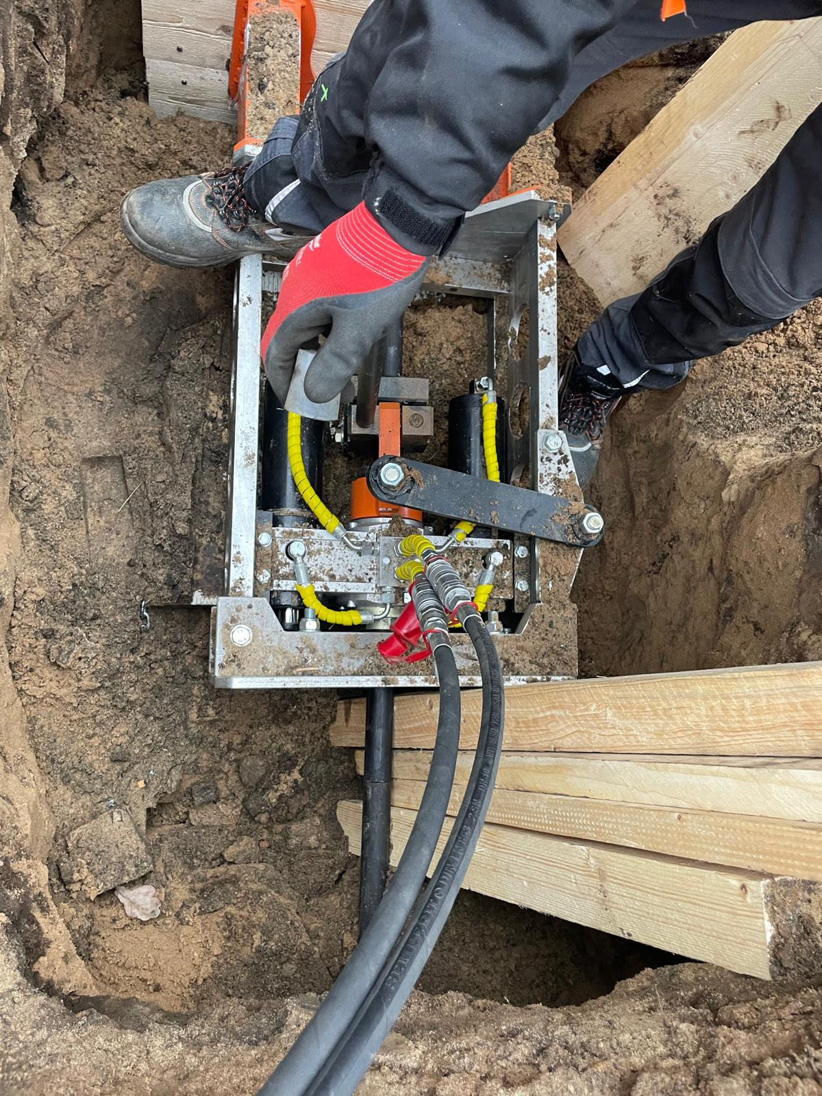 Widok z góry na urządzenie do robót ziemnych w wykopie, z widocznymi elementami hydraulicznymi, w trakcie pracy, z pracownikiem w rękawicach ochronnych i butach roboczych