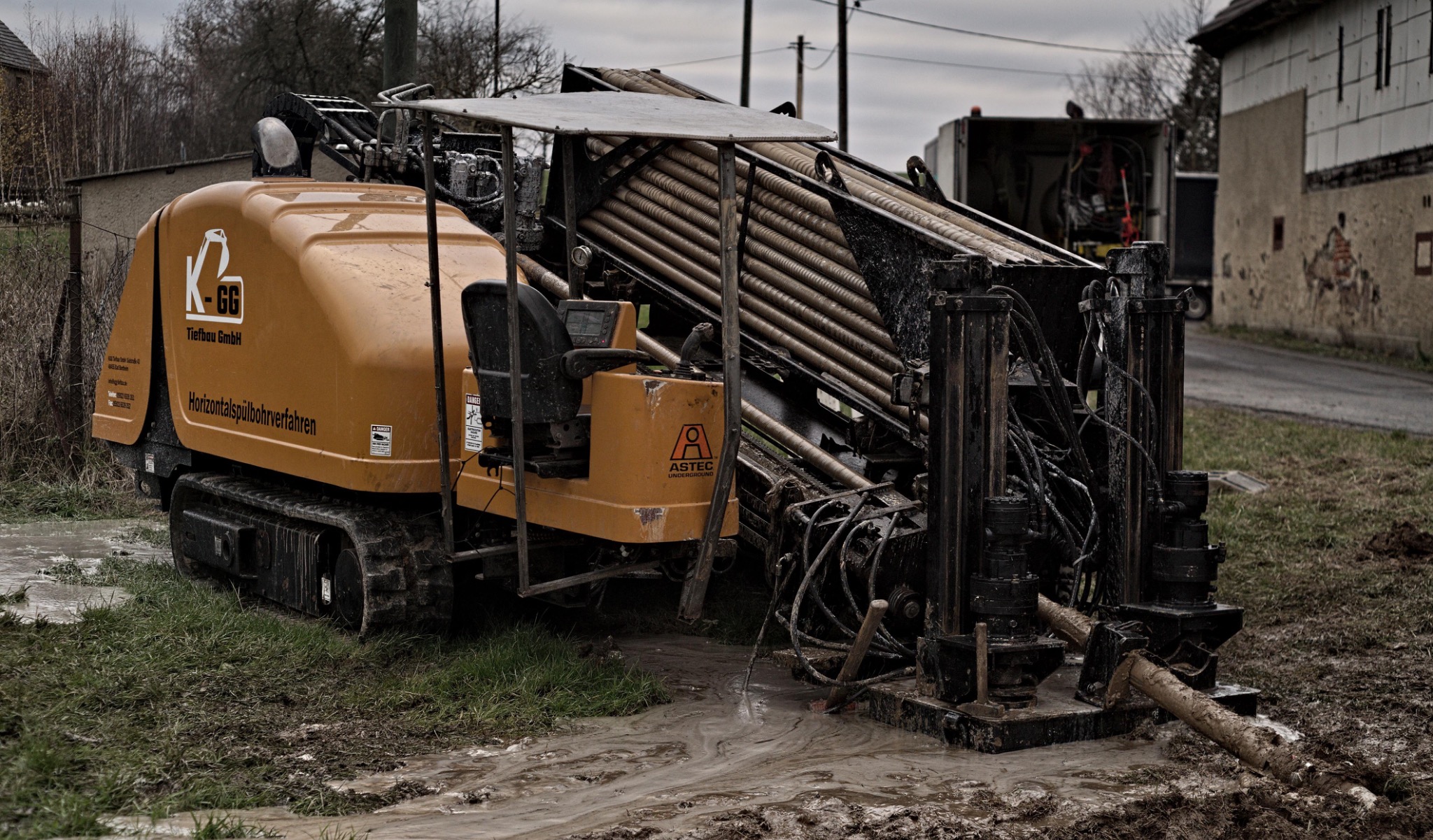 Pomarańczowa maszyna do robót ziemnych z gąsienicami i rurami w błotnistym terenie, widoczny fragment budynku i samochód w tle.