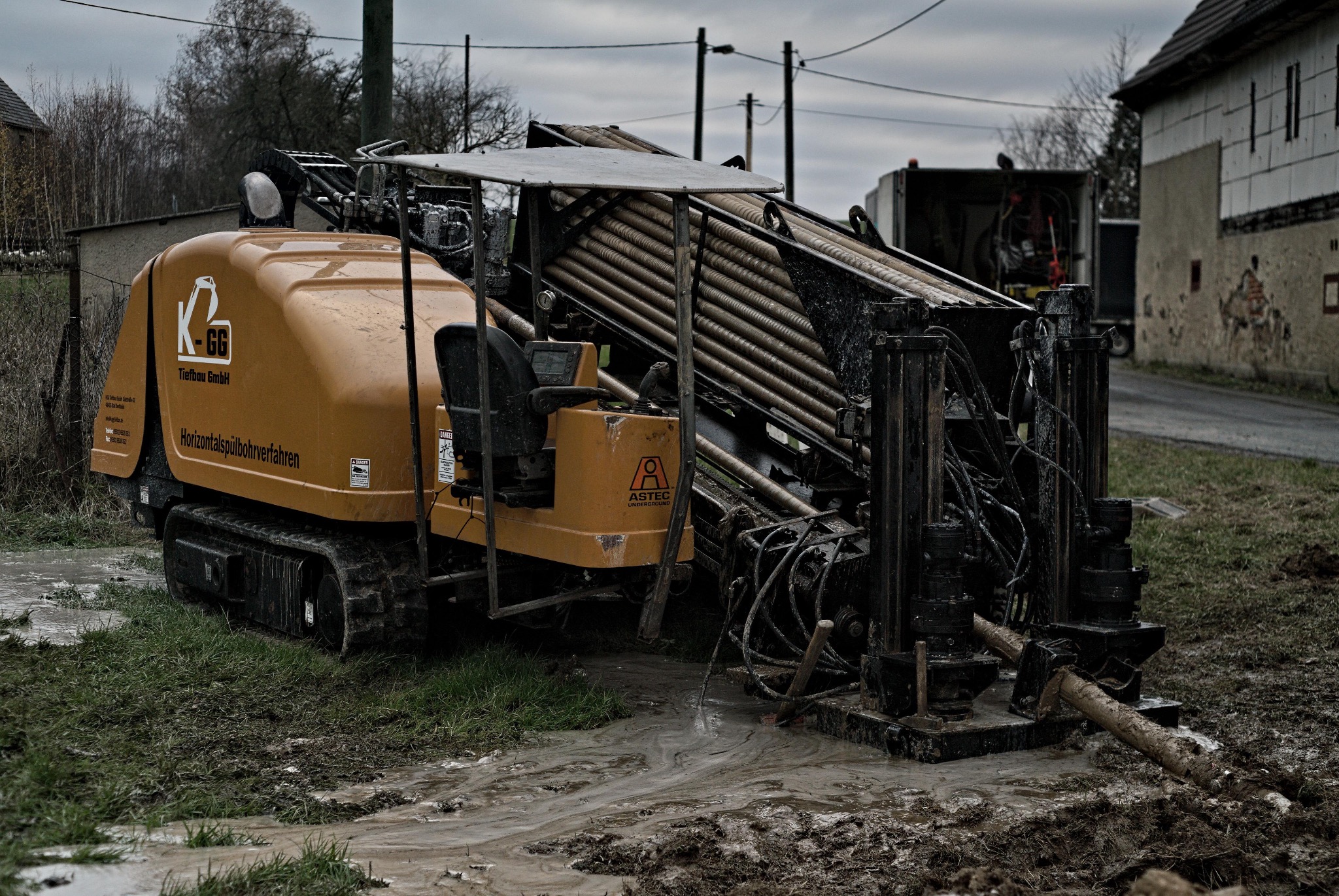 Żółta maszyna do wiercenia horyzontalnego na gąsienicach, z widocznymi rurami w podajniku, stoi w błotnistym terenie obok drogi i budynków.