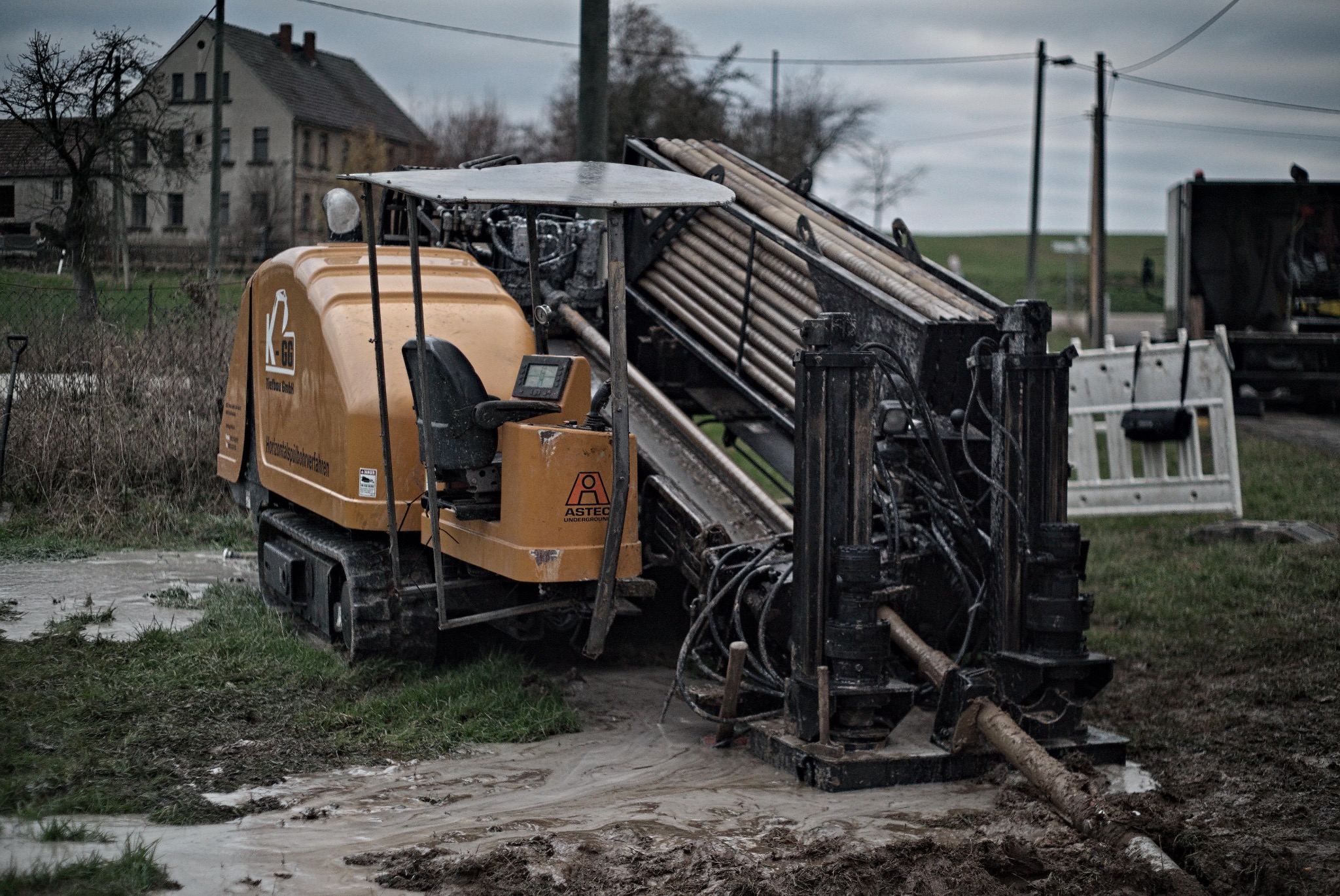 Żółta maszyna do wiercenia poziomego z rurami na błotnistym terenie, w tle budynek mieszkalny i pochmurne niebo.