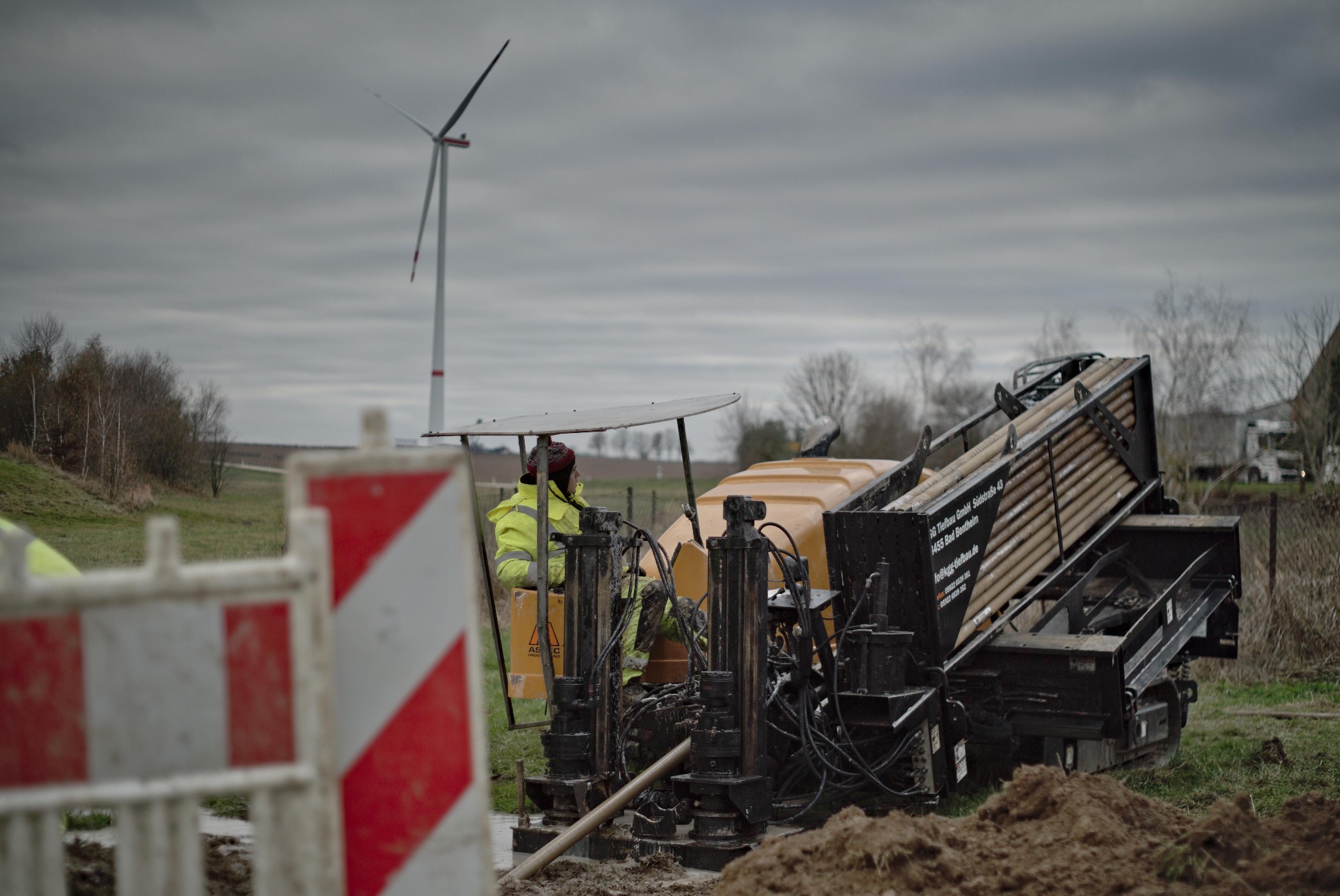 Operator w żółtej kurtce obsługuje maszynę do wiercenia horyzontalnego na tle krajobrazu z turbiną wiatrową, widoczne zabezpieczenia budowy.