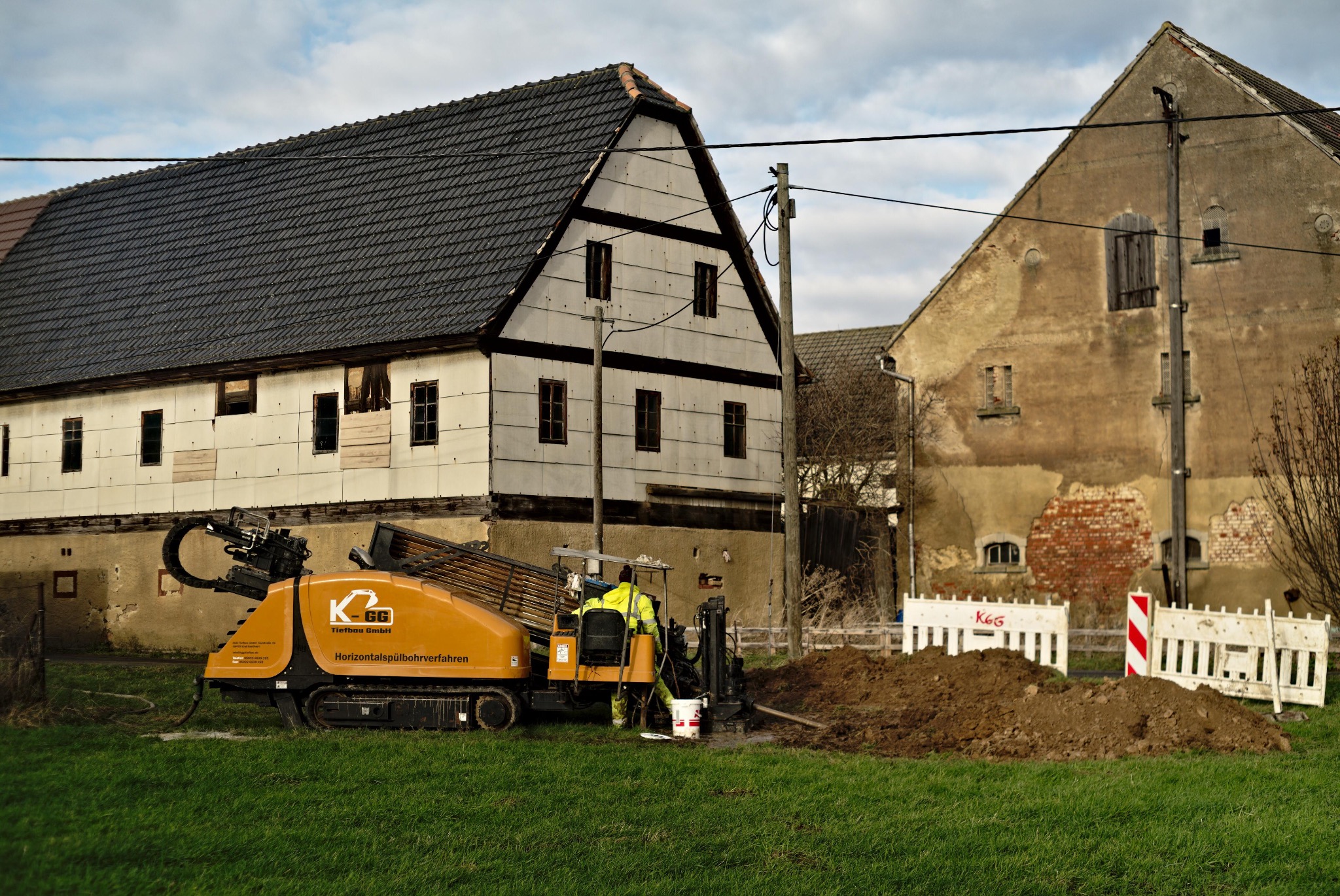 Żółta maszyna do robót ziemnych z operatorem w kamizelce odblaskowej na tle starych budynków gospodarczych i sterty ziemi.