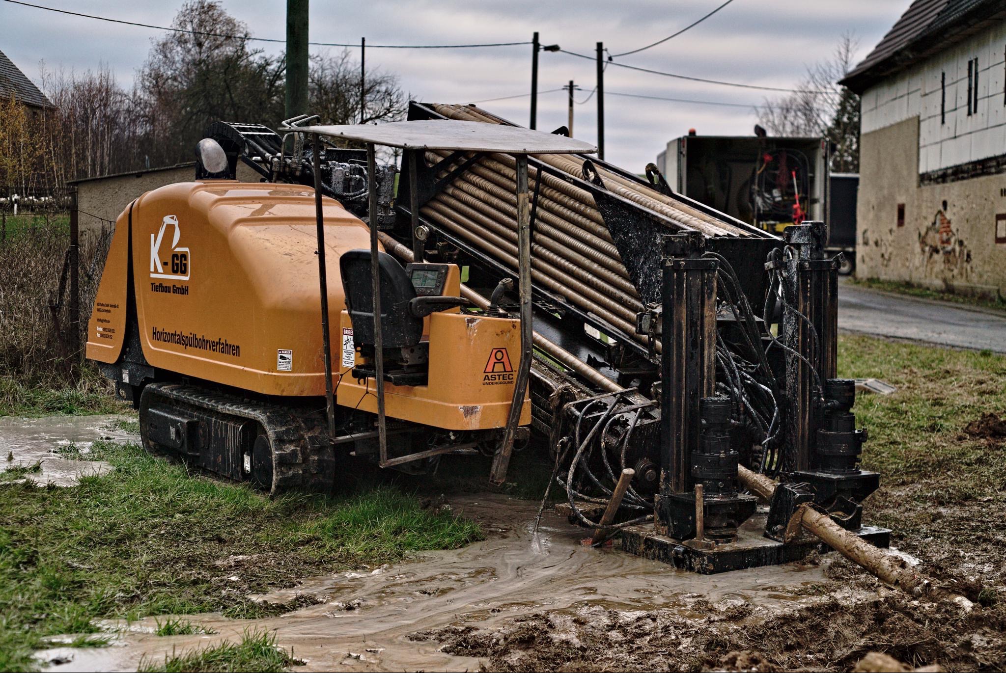 Pomarańczowa maszyna do przewiertów sterowanych firmy Astec, z widocznymi rurami w koszu, stoi w błotnistym terenie na tle wiejskiej zabudowy w pochmurny dzień.