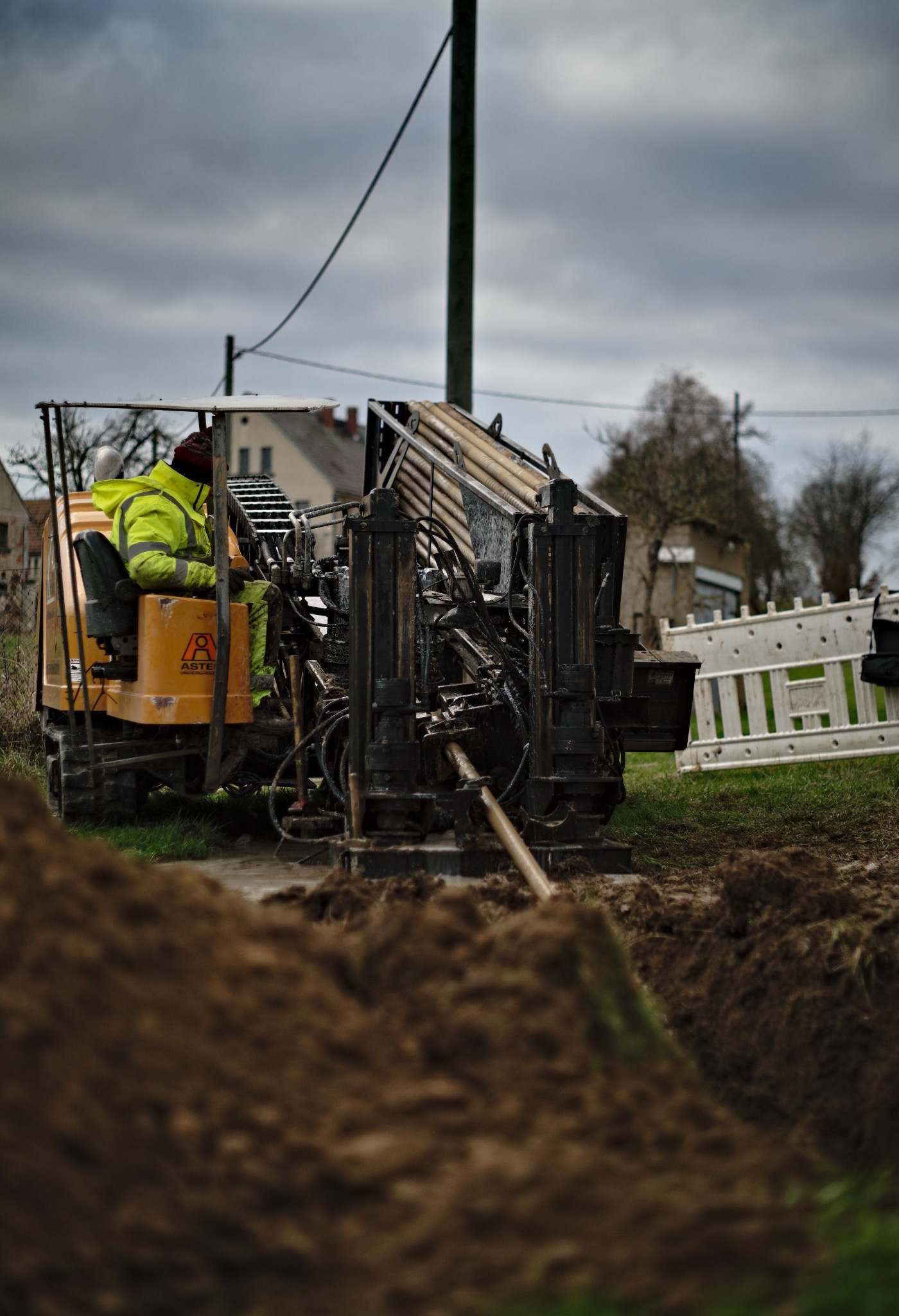 Operator w jaskrawej kurtce obsługuje maszynę do wiercenia horyzontalnego, widoczna rura w ziemi, w tle zabudowania mieszkalne i pochmurne niebo.