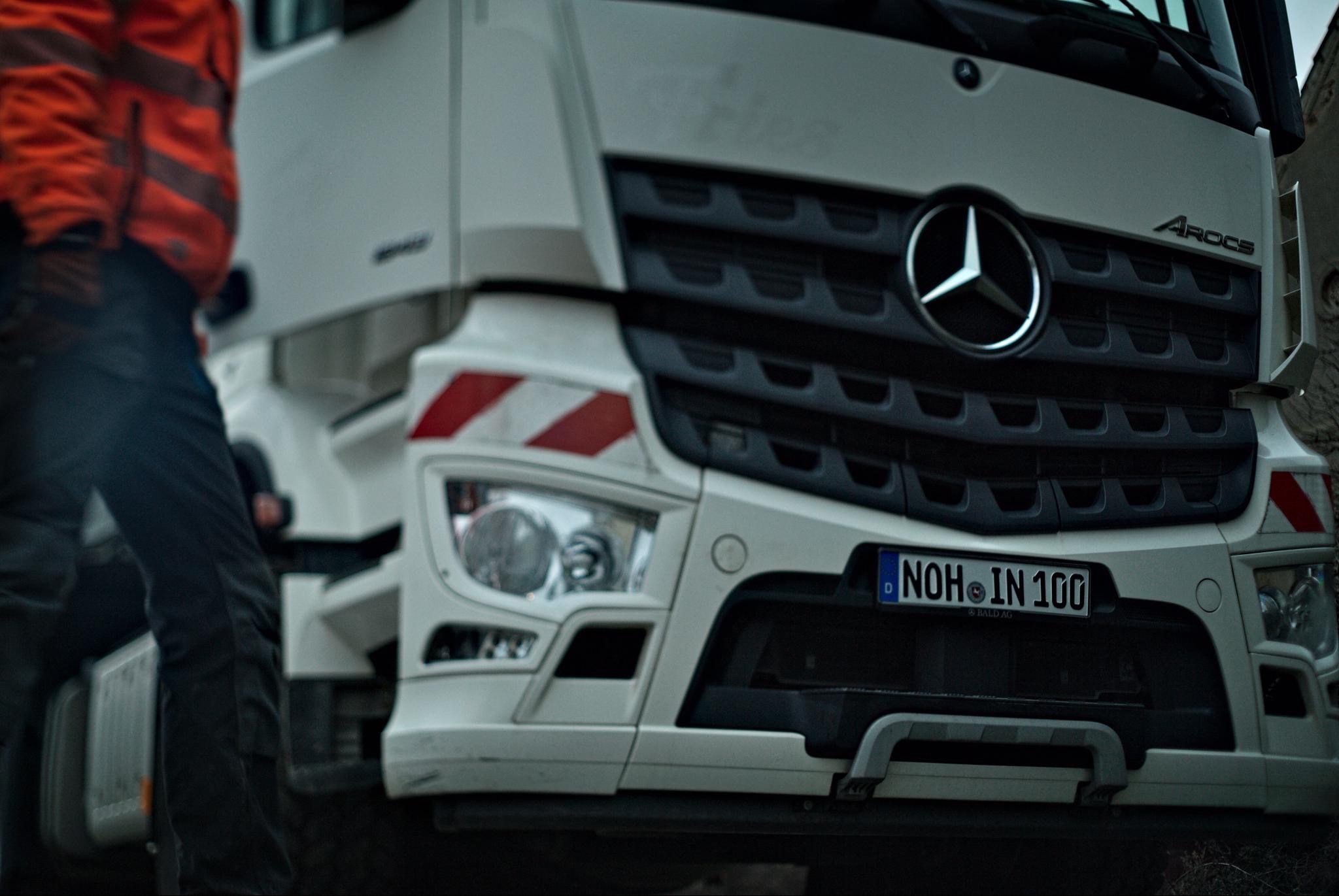 Przód białej ciężarówki Mercedes-Benz Arocs z widocznym logo i tablicą rejestracyjną NOH IN 100, częściowo zasłonięty przez osobę w pomarańczowej odzieży roboczej.