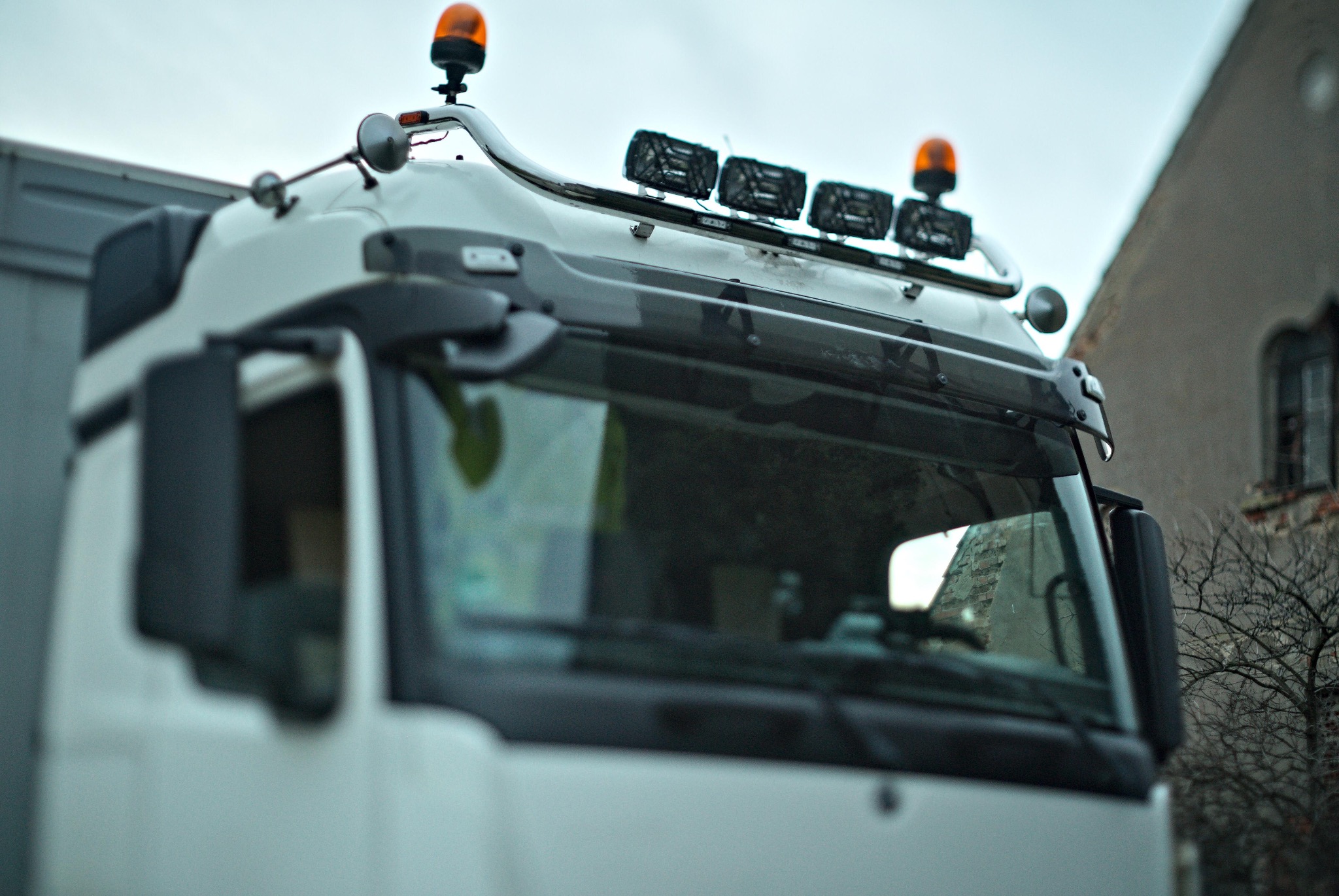 Kabina białego samochodu ciężarowego z pomarańczowymi światłami ostrzegawczymi i dodatkowym oświetleniem, widziana z dołu na tle budynku w Kołobrzegu.