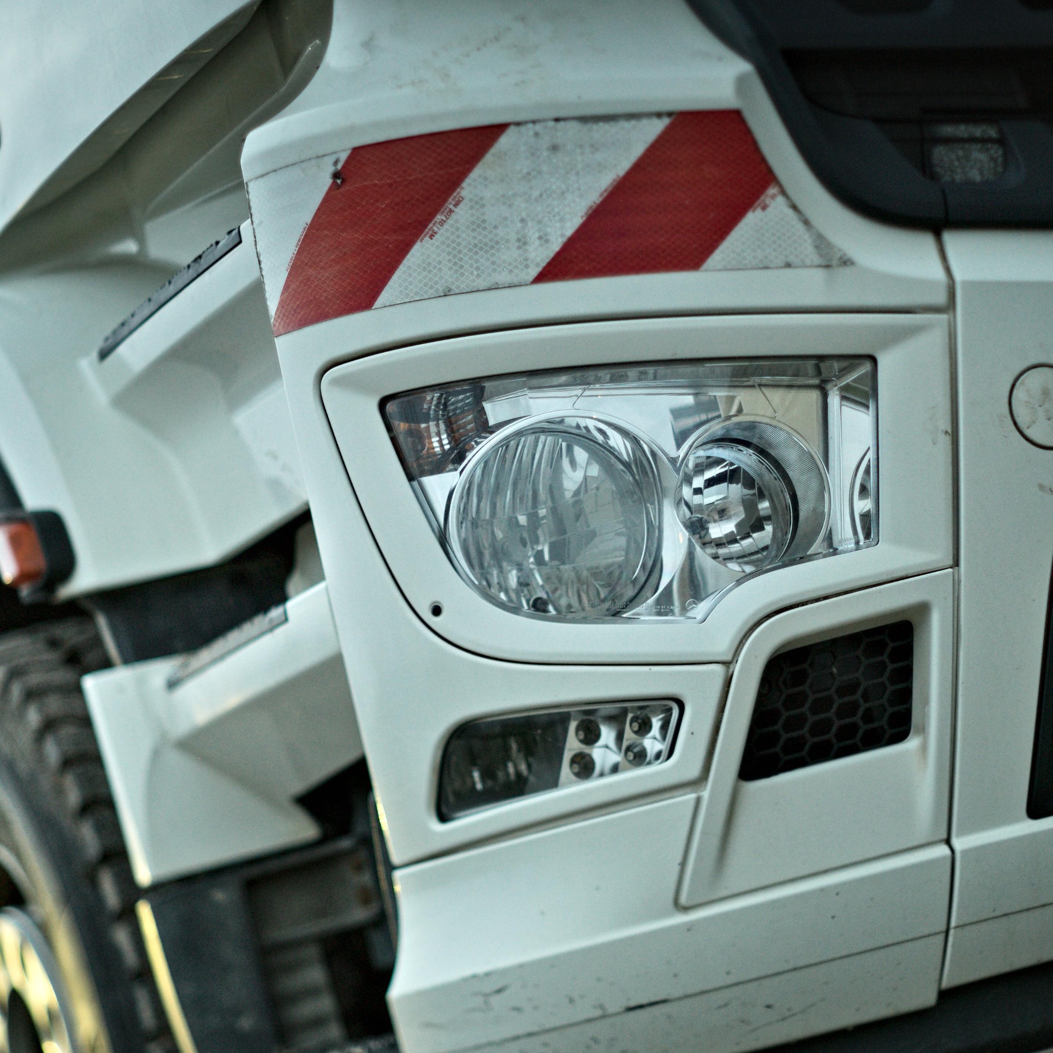 Fragment białej ciężarówki z widocznym reflektorem i odblaskowymi pasami ostrzegawczymi.