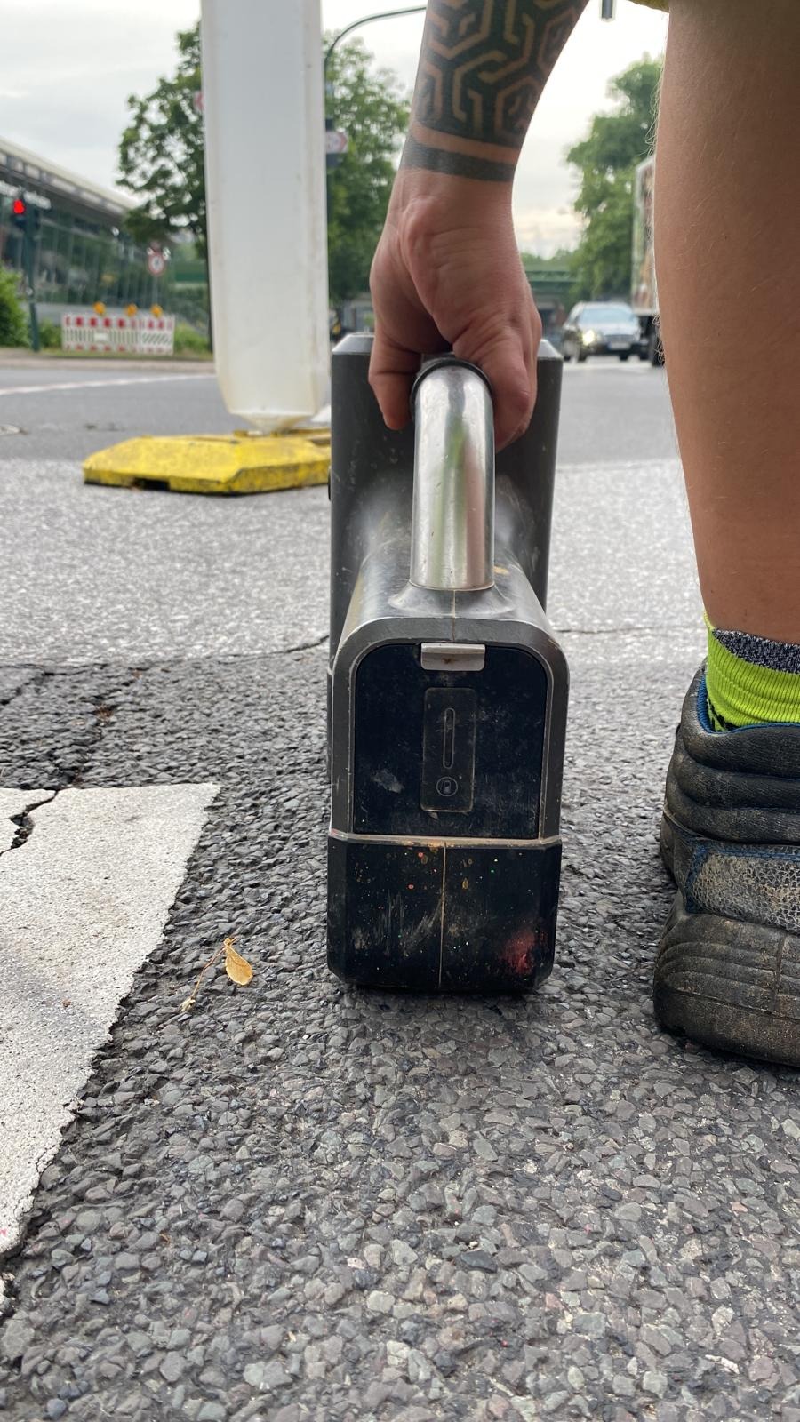 Przenośne urządzenie pomiarowe trzymane w dłoni z tatuażem, obok but roboczy i żółta skarpetka, na tle ulicy