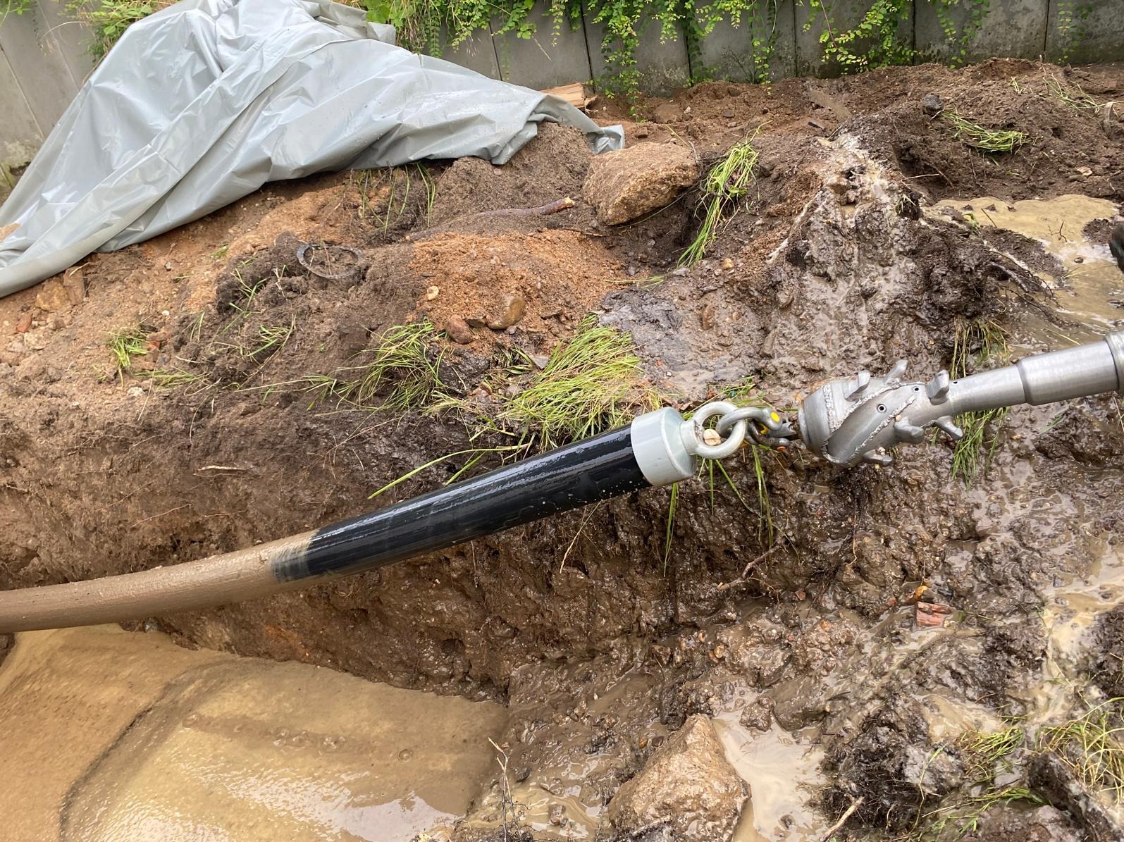 Wiertnica z głowicą frezującą w wykopie wypełnionym błotem, widoczny fragment rury i łańcuch łączący elementy.