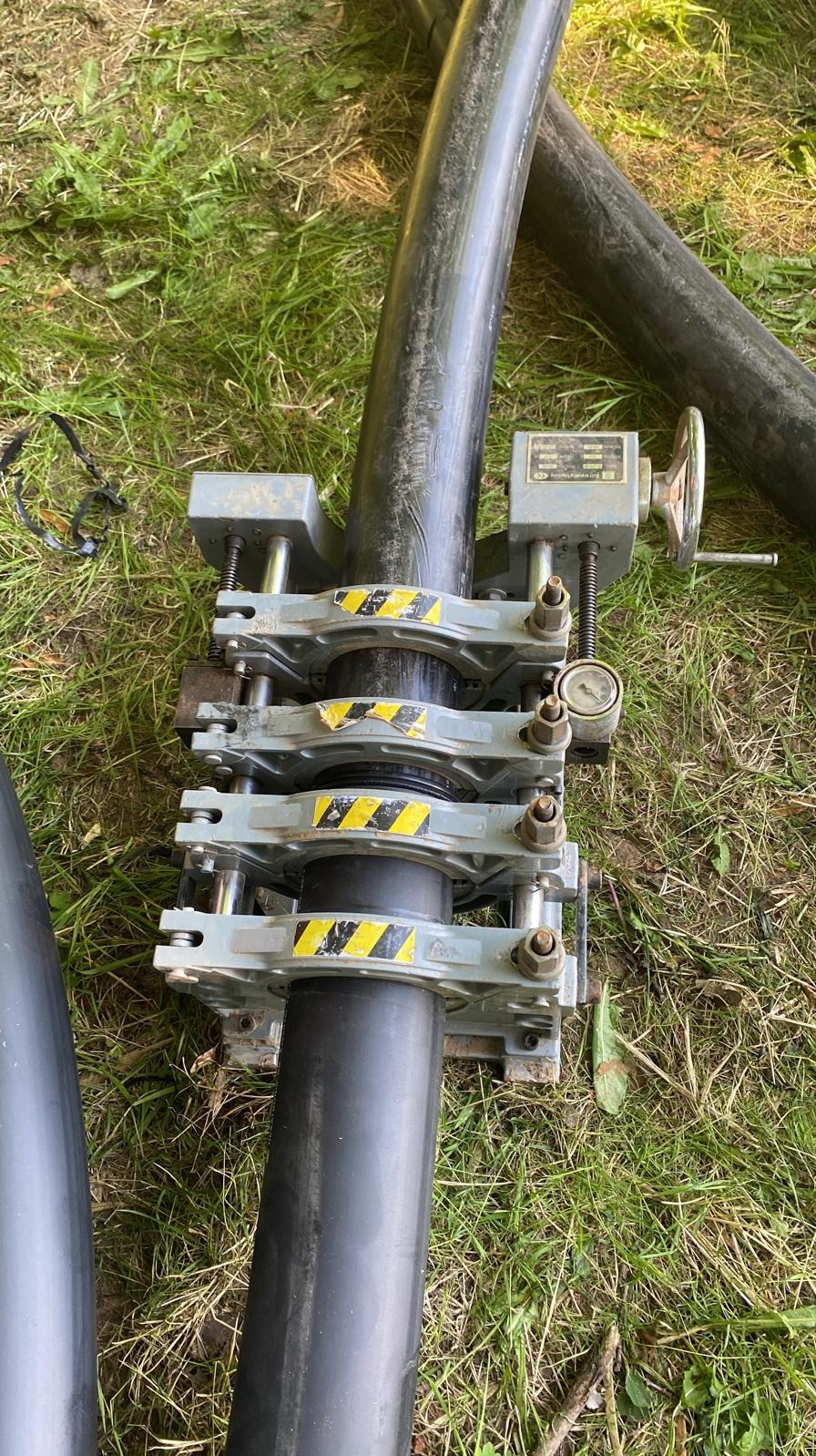 Zgrzewanie rur HDPE przy pomocy zgrzewarki doczołowej na trawie, widoczne czarne rury, złączki z żółto-czarnymi pasami ostrzegawczymi, manometr i pokrętło regulacyjne.