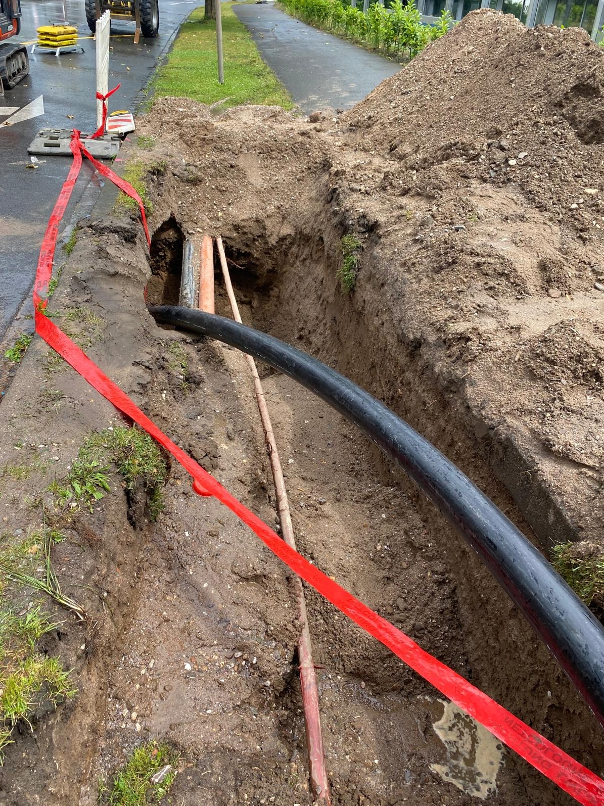 Wykop w ziemi z widocznymi rurami: czarną, szarą i pomarańczową; zabezpieczony czerwoną taśmą ostrzegawczą, na tle zieleni i chodnika.