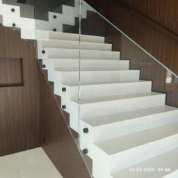 Vasyl Stetsiuk - Nowoczesne białe schody z szklaną balustradą i drewnianym wykończeniem ściany. Minimalistyczny design z czarnymi elementami mocującymi szkło.