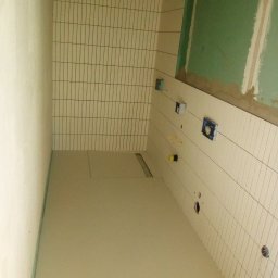 Vasyl Stetsiuk - Wnętrze łazienki w trakcie prac: ułożone wąskie, pionowe płytki na ścianach, podłoga wylewka, widoczne otwory pod instalacje i fragment ściany z zieloną płytą.
