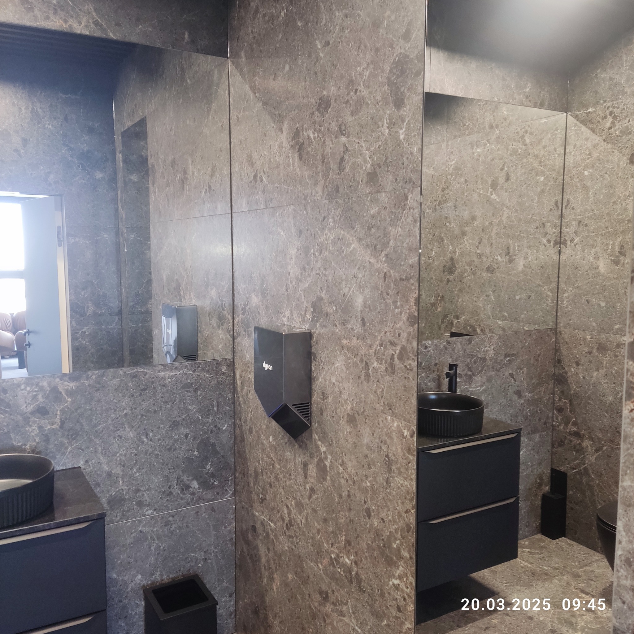 Nowoczesna łazienka z szarymi płytkami imitującymi marmur, czarną umywalką nablatową i podajnikiem do rąk Dyson. Lustra powiększają przestrzeń.