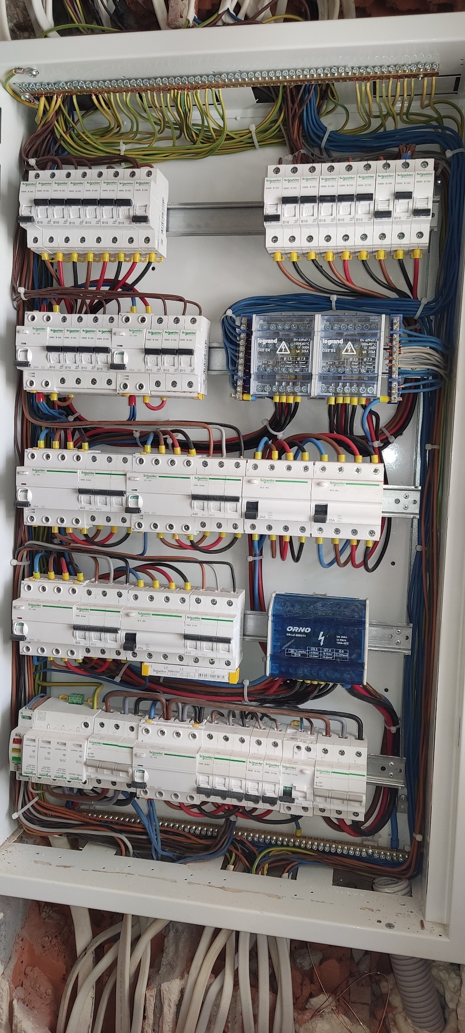Rozdzielnica elektryczna z zamontowanymi bezpiecznikami i przewodami. Widoczne kable w różnych kolorach, aparatura modułowa firm Schneider i Legrand oraz listwa zaciskowa.