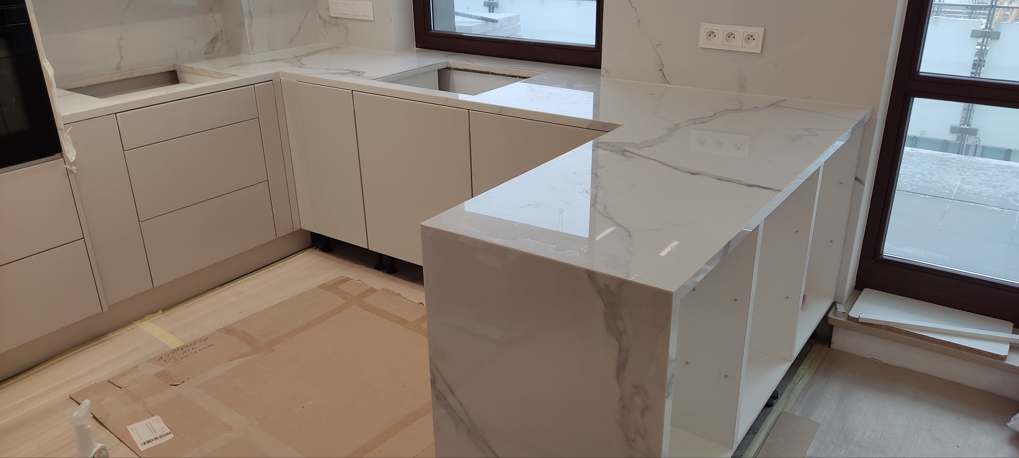 Nowoczesna, jasna kuchnia z marmurowym blatem i białymi szafkami, widok z góry, zabezpieczona podłoga kartonem, okno w tle.