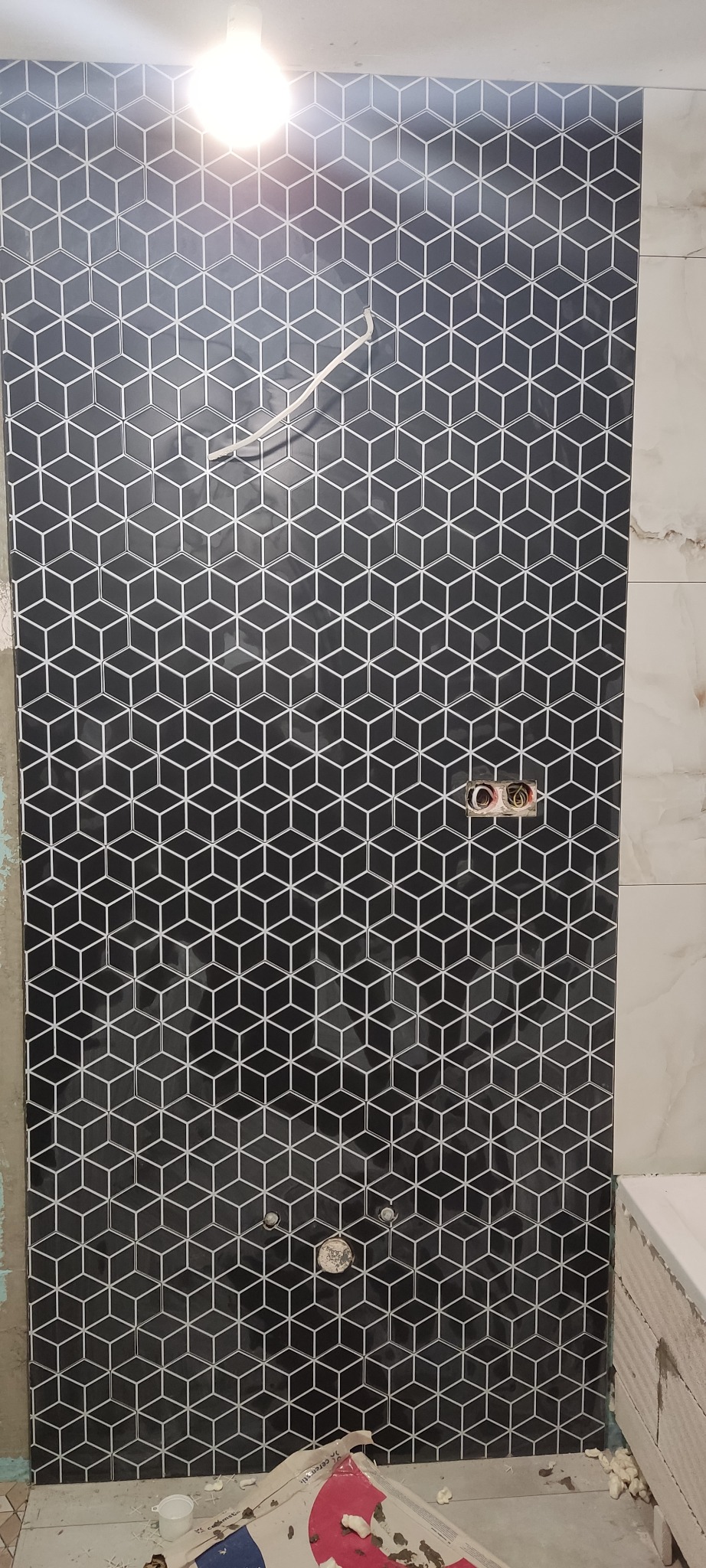 Ściana łazienki wyłożona czarnymi, błyszczącymi płytkami z białym geometrycznym wzorem kostek 3D. Widoczne gniazdka elektryczne i fragment wanny.