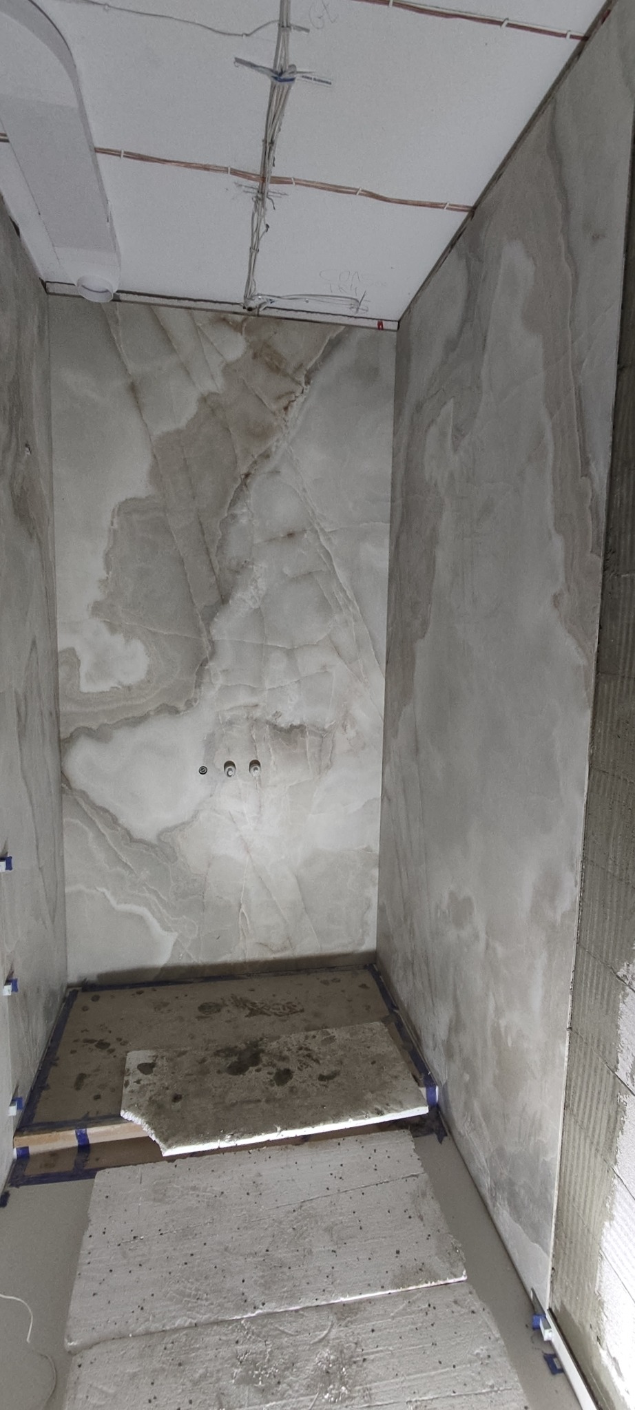 Wnętrze łazienki w trakcie prac. Położone płytki imitujące marmur na ścianach, widoczne kable pod sufitem, częściowo ułożona podłoga.