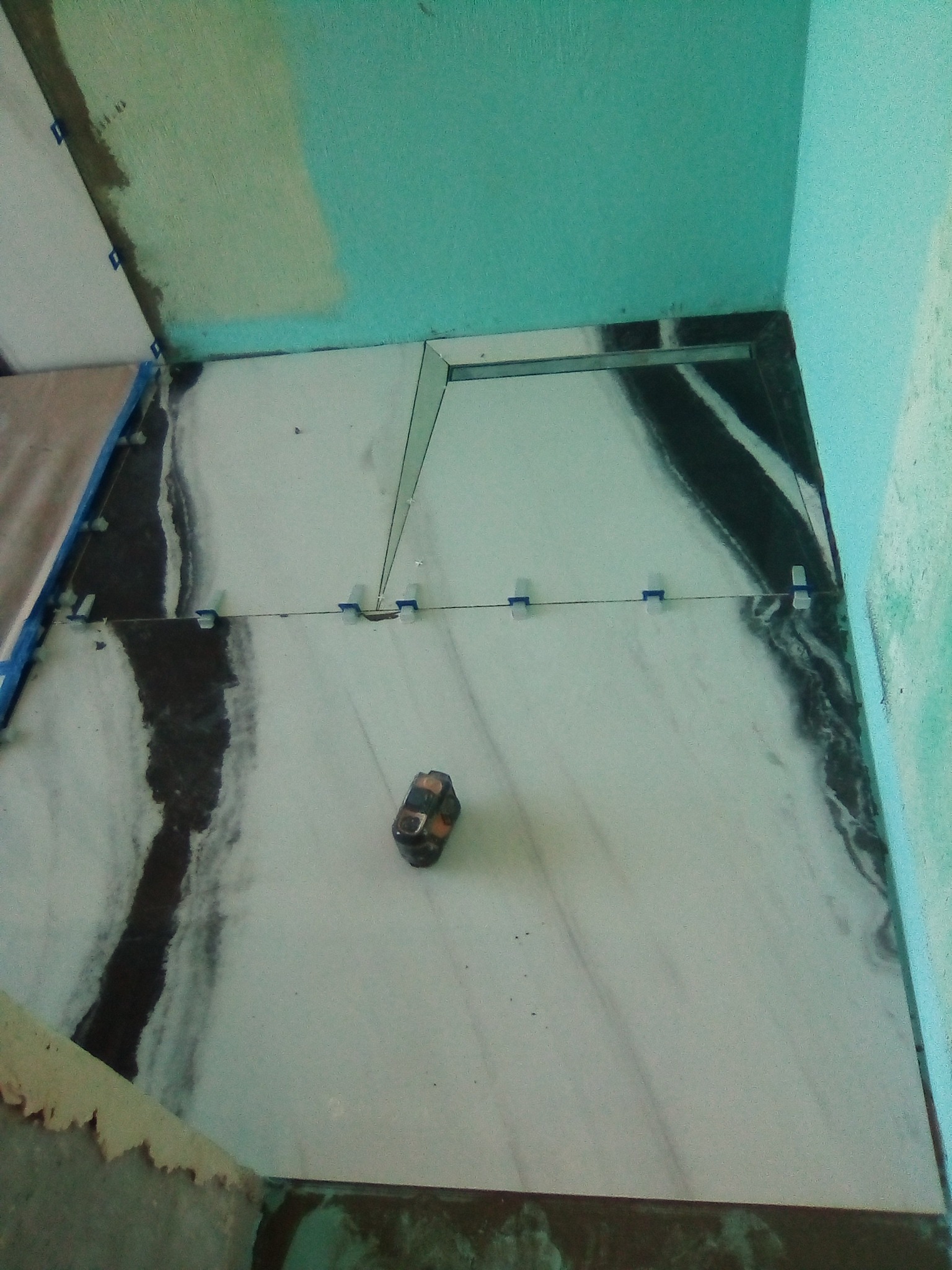 Fragment łazienki w trakcie prac: ułożone płytki imitujące marmur z fugami i poziomnicą. Widoczne niebieskie ściany i elementy mocujące płytki.