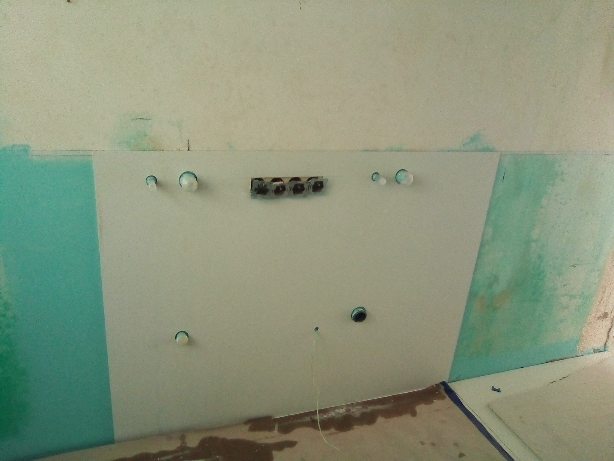 Ściana z przygotowanymi otworami pod instalacje elektryczne i wodne, częściowo pomalowana na turkusowo, w trakcie prac wykończeniowych. Widoczne przewody.