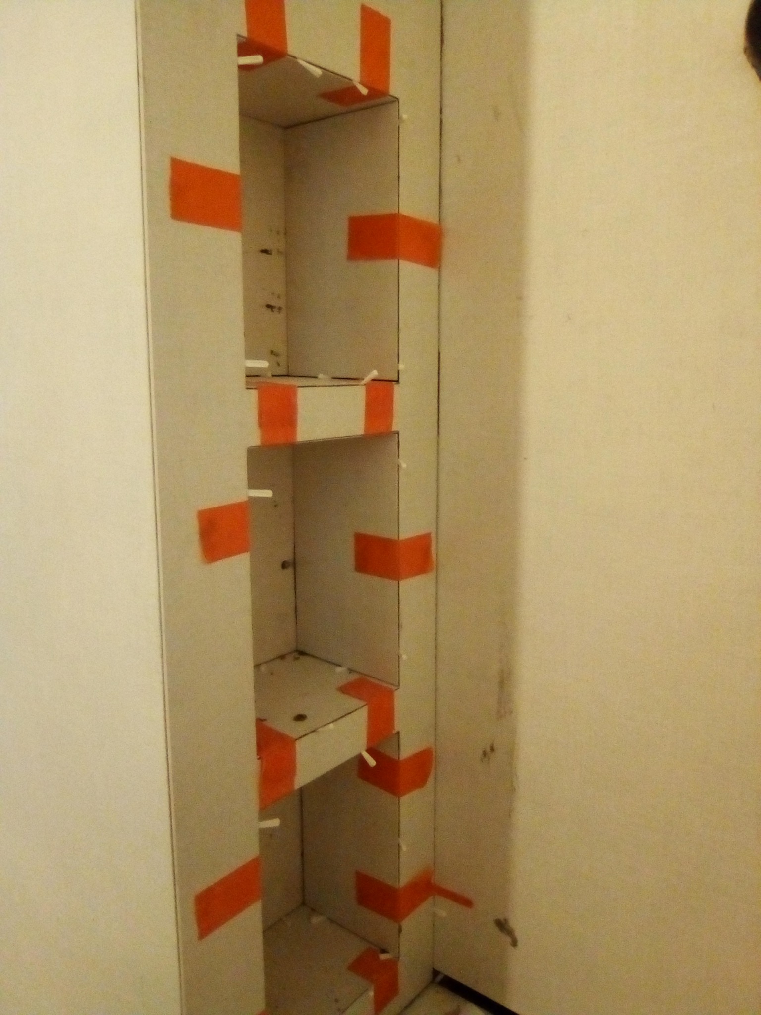 Narożna wnęka w ścianie, wyłożona płytkami, zabezpieczona pomarańczową taśmą. Widoczne białe krzyżyki dystansowe. Etap prac wykończeniowych.