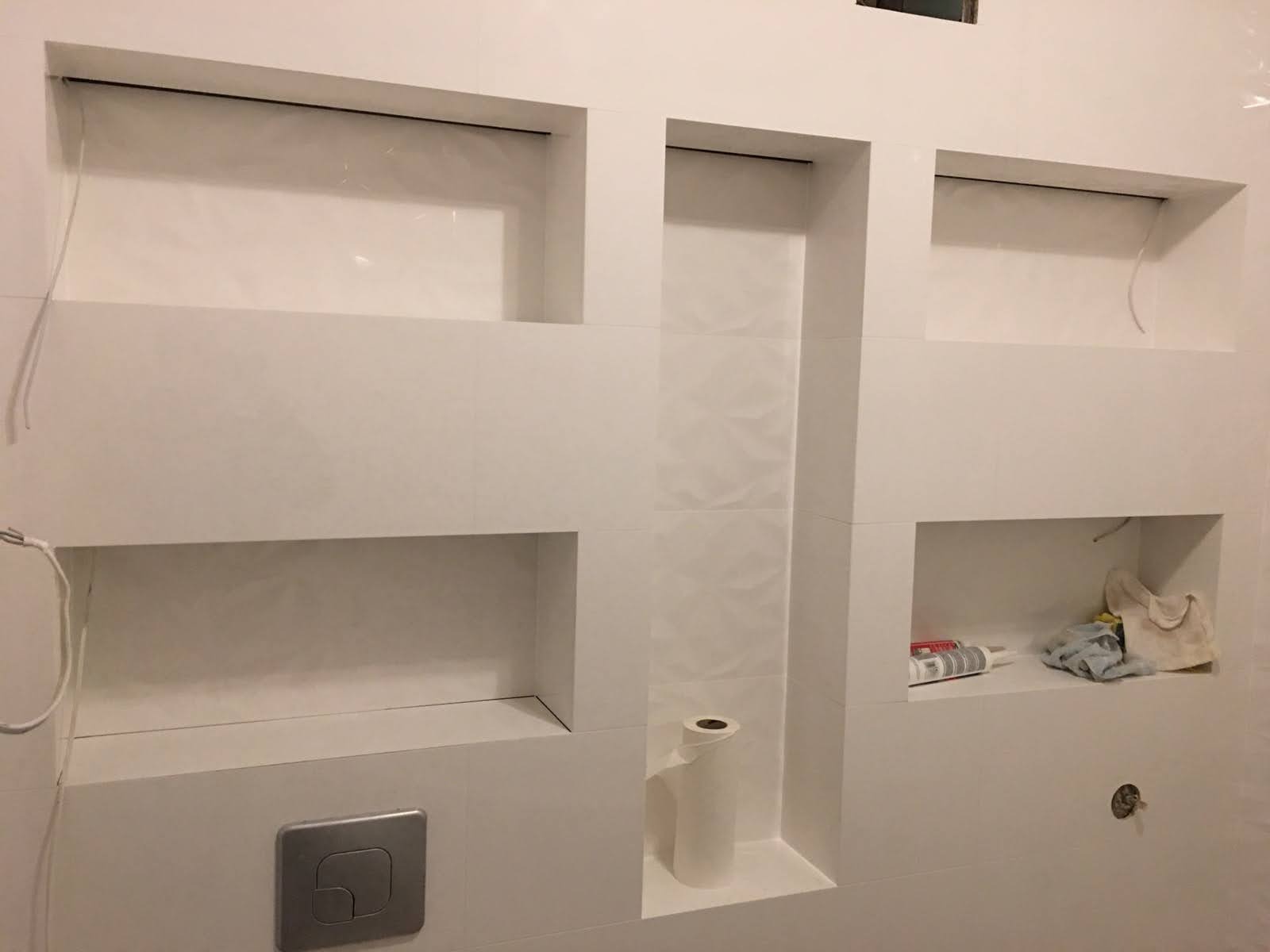 Biała łazienka z wnękami wyłożonymi płytkami, widoczny przycisk spłuczki, rolka papieru toaletowego i akcesoria do montażu, detale wykończenia.