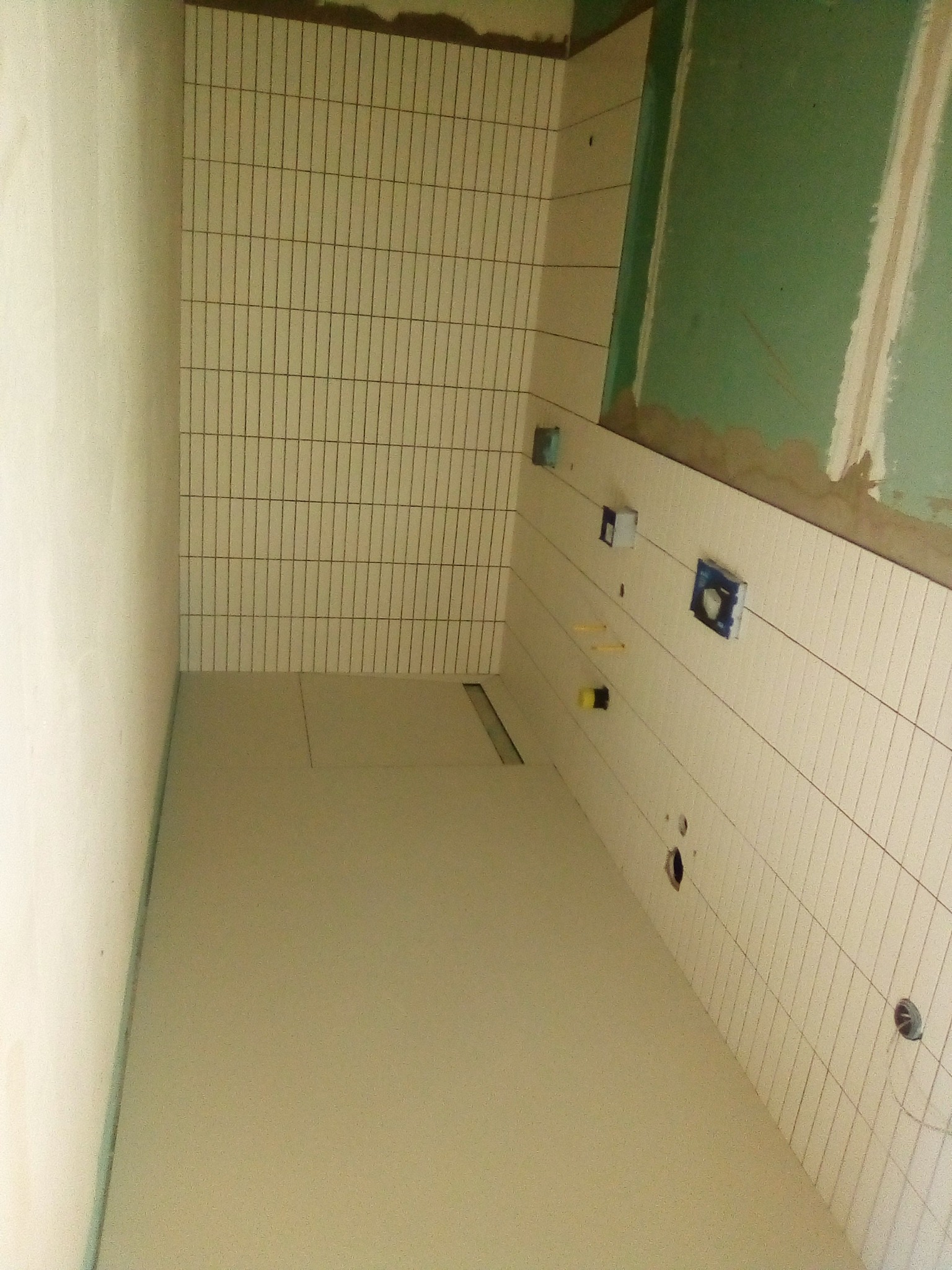 Wnętrze łazienki w trakcie prac: ułożone wąskie, pionowe płytki na ścianach, podłoga wylewka, widoczne otwory pod instalacje i fragment ściany z zieloną płytą.