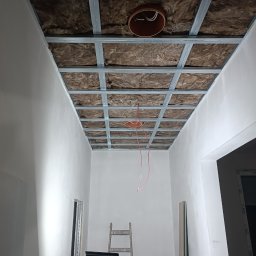 GLANZ - Sufit podwieszany w trakcie montażu: metalowa konstrukcja, izolacja z wełny mineralnej, wystające przewody elektryczne. Widoczna drabina i materiały budowlane.