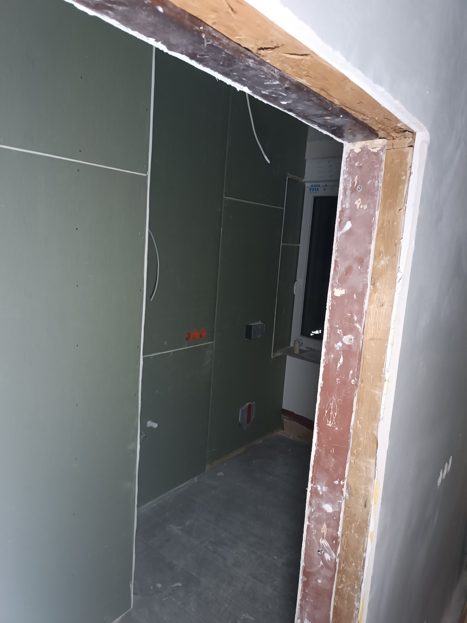 Wnętrze w trakcie prac: ściany wyłożone płytami gipsowo-kartonowymi, widoczne instalacje elektryczne i fragment okna przez surową futrynę drzwiową.