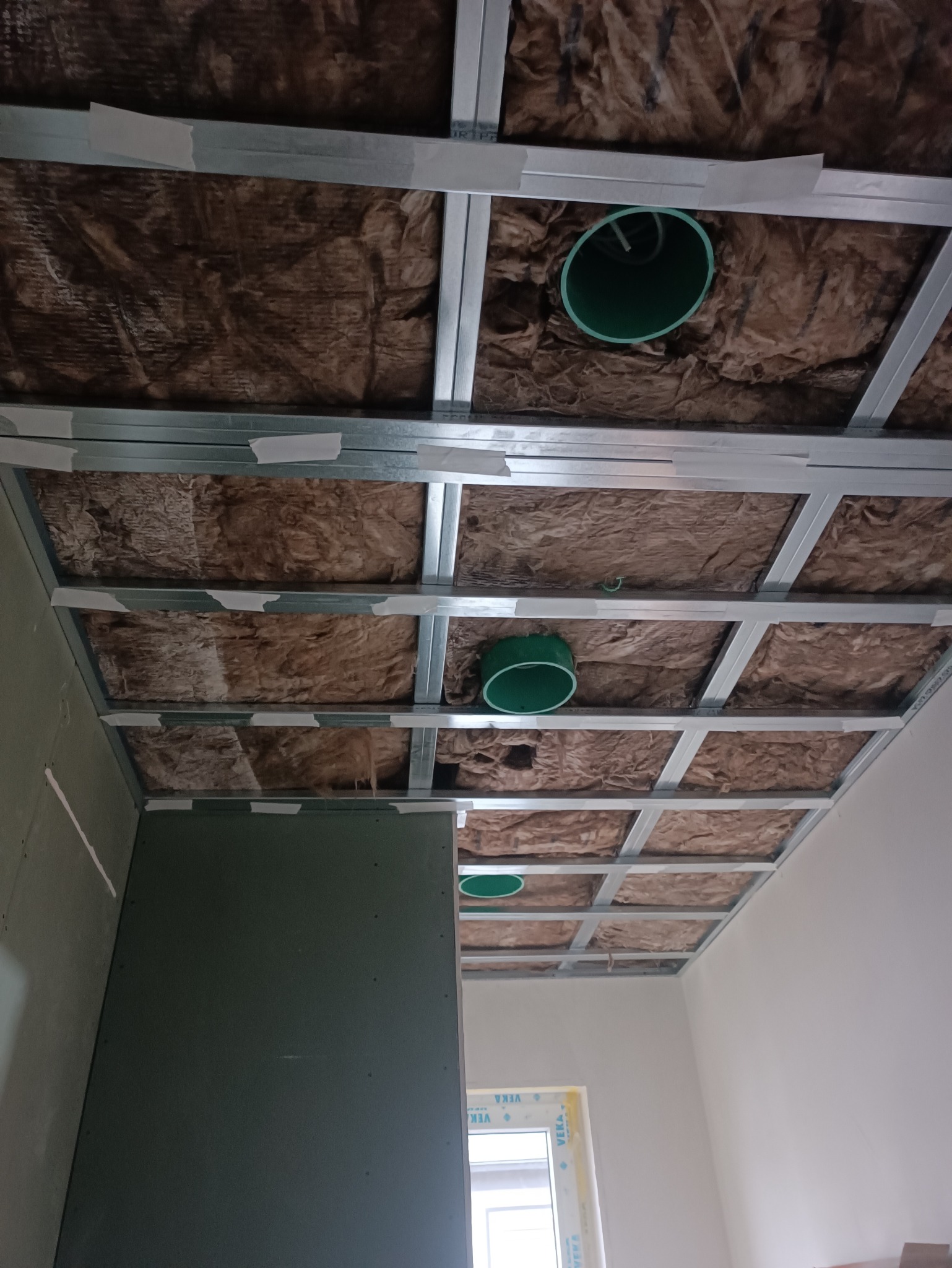 Sufit podwieszany z izolacją z wełny mineralnej, widoczne profile metalowe i otwory wentylacyjne. Częściowo zamontowana płyta gipsowo-kartonowa.