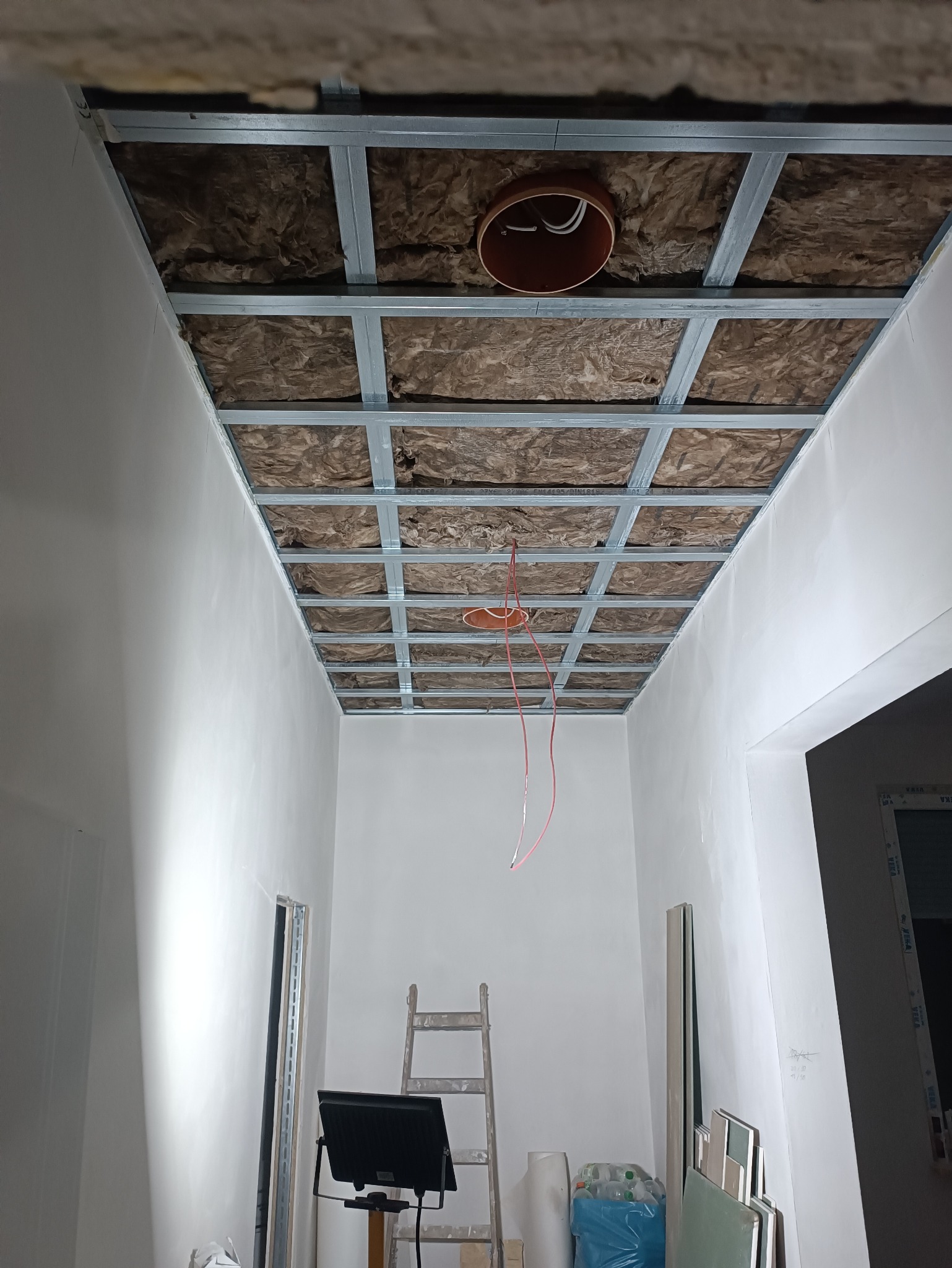 Sufit podwieszany w trakcie montażu: metalowa konstrukcja, izolacja z wełny mineralnej, wystające przewody elektryczne. Widoczna drabina i materiały budowlane.
