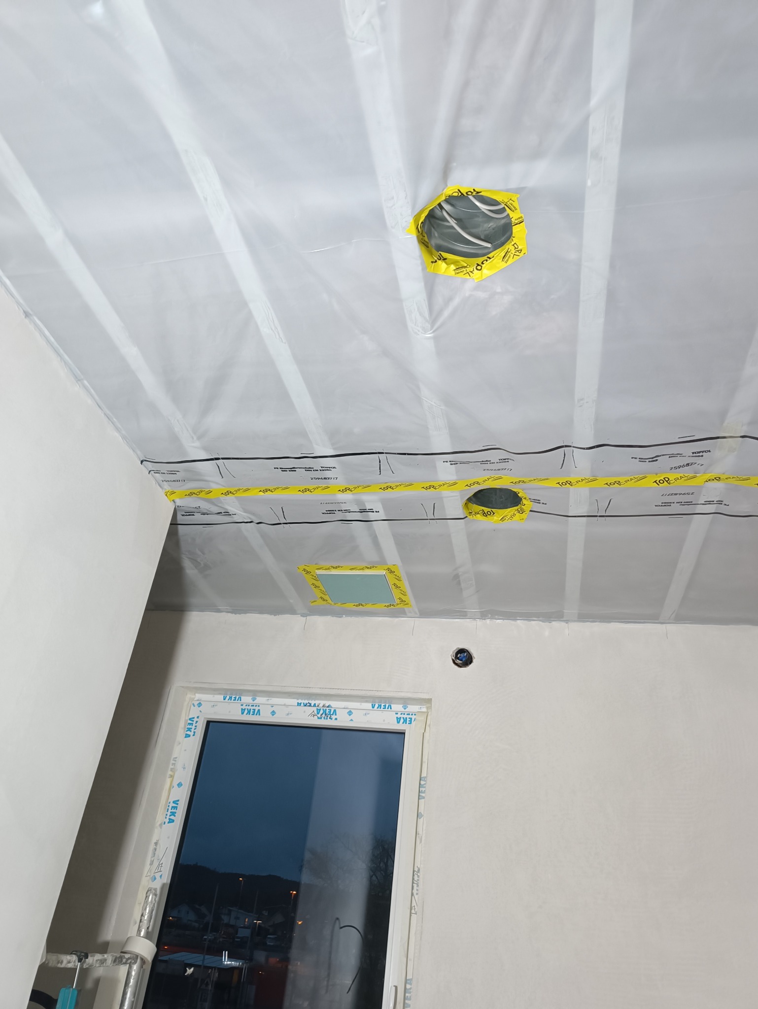 Przygotowanie do malowania: sufit i ściany zabezpieczone folią ochronną z taśmą, widoczne otwory na oświetlenie. Okno z PCV. Prace wykończeniowe w trakcie.