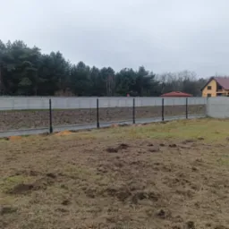 Montaż ogrodzenia panelowego z podmurówką betonową i czarnymi słupkami na nierównym terenie, w tle las i dom z czerwoną dachówką.