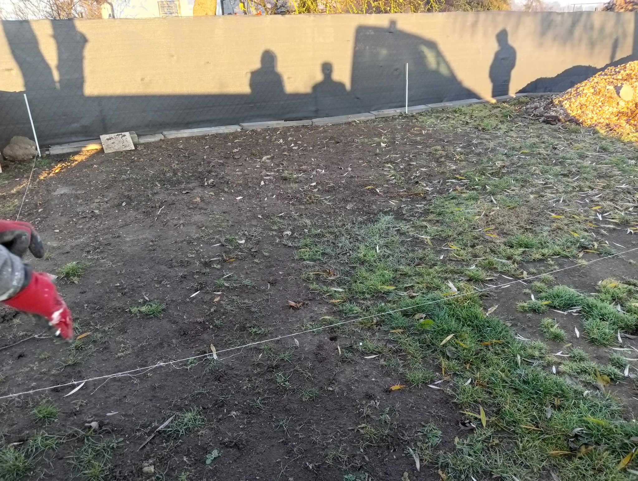 Przygotowanie terenu pod ogrodzenie: wytyczone sznurkiem granice, widoczne słupki i cień osób na tymczasowej siatce osłonowej, pryzma liści w rogu działki.