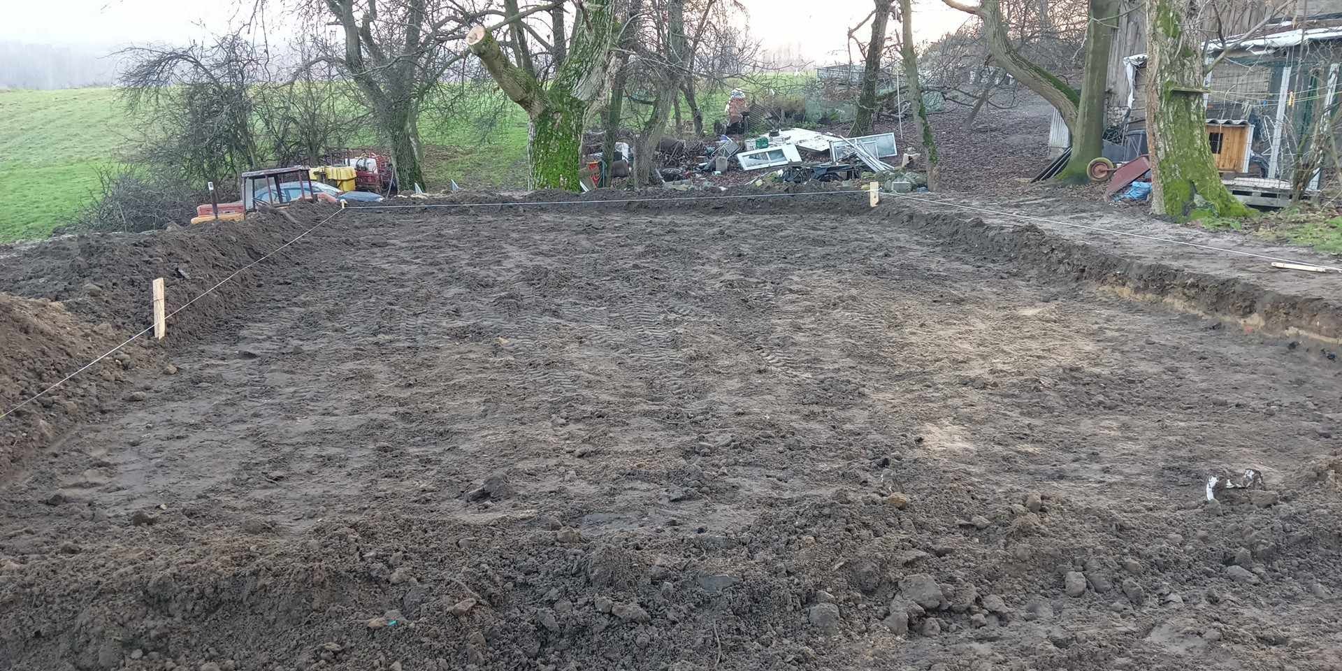 Przygotowany teren pod budowę, widoczne wytyczone granice za pomocą sznurka i kołków, ziemia wyrównana, w tle drzewa i sprzęt budowlany.