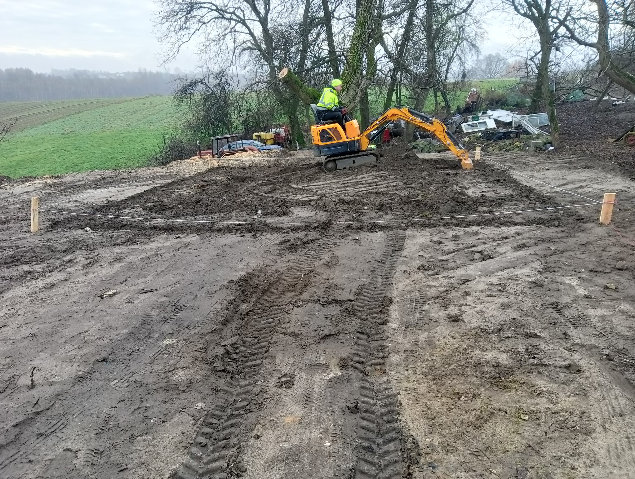 Pomarańczowa mini koparka z operatorem w żółtej kamizelce podczas prac ziemnych na błotnistym terenie, wyznaczone drewniane słupki z linką, w tle drzewa i zielone pole.