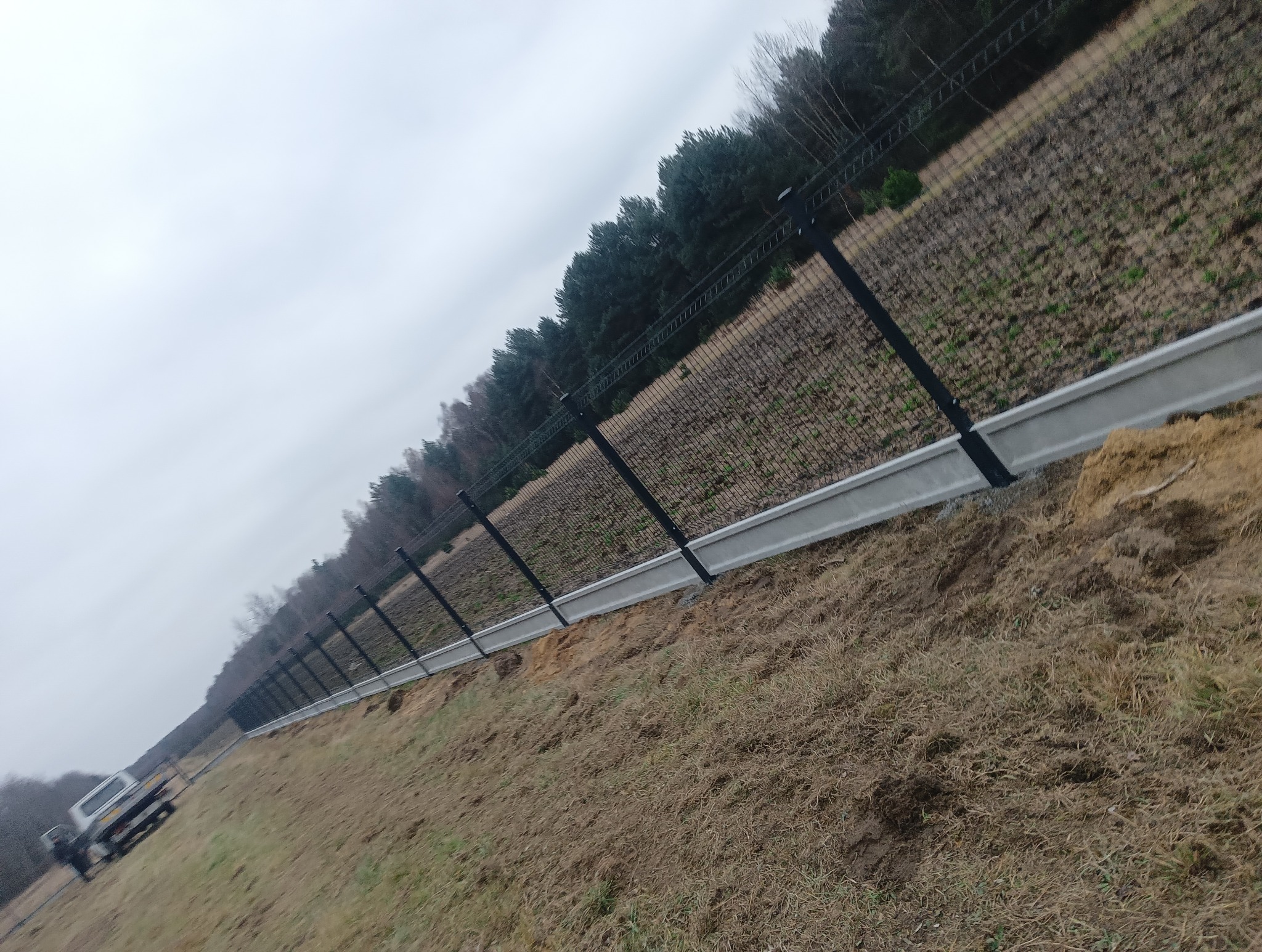 Długi, czarny system ogrodzeniowy z siatki na betonowej podmurówce, rozciągający się wzdłuż nierównego terenu z widocznym lasem w tle i samochodem dostawczym w oddali.