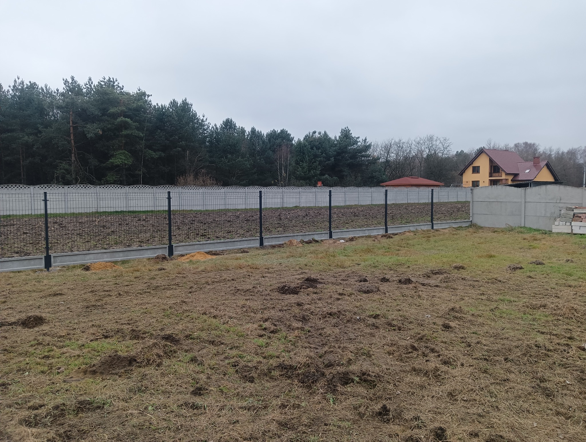 Montaż ogrodzenia panelowego z podmurówką betonową i czarnymi słupkami na nierównym terenie, w tle las i dom z czerwoną dachówką.