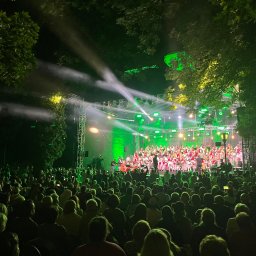 btone.tech sp. z o.o. - Oświetlona zielonymi światłami scena plenerowa z występem chóru i orkiestry w strojach ludowych, oglądana przez duży tłum publiczności.