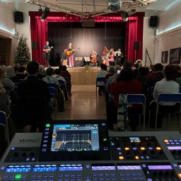 btone.tech sp. z o.o. - Koncert z udziałem kilku muzyków grających na instrumentach, widok zza stołu mikserskiego z widoczną publicznością i choinką w tle.