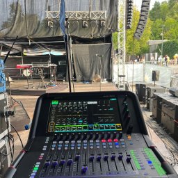 btone.tech sp. z o.o. - Konsoleta mikserska Allen & Heath Avantis Solo na tle sceny plenerowej z nagłośnieniem liniowym i instrumentami muzycznymi, widok zza urządzenia.