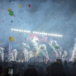btone.tech sp. z o.o. - Koncert z tłumem, scena oświetlona reflektorami, konfetti w powietrzu i dym z wytwornic. W tle konstrukcja sceniczna i telebimy. Widok zza publiczności.