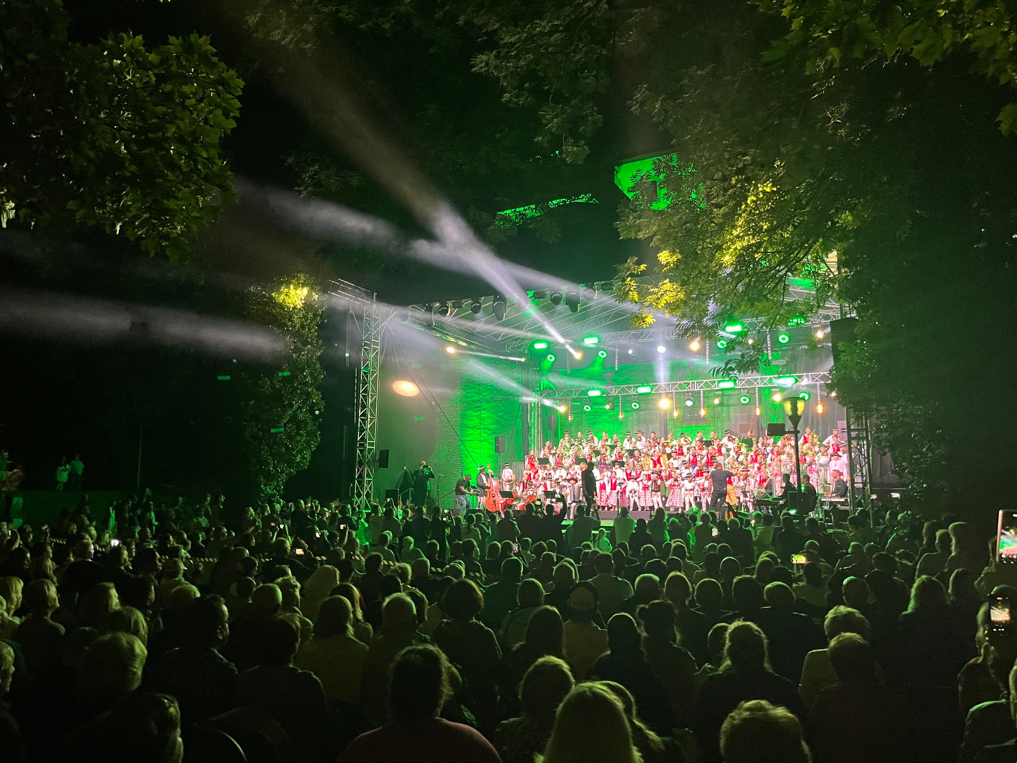Oświetlona zielonymi światłami scena plenerowa z występem chóru i orkiestry w strojach ludowych, oglądana przez duży tłum publiczności.