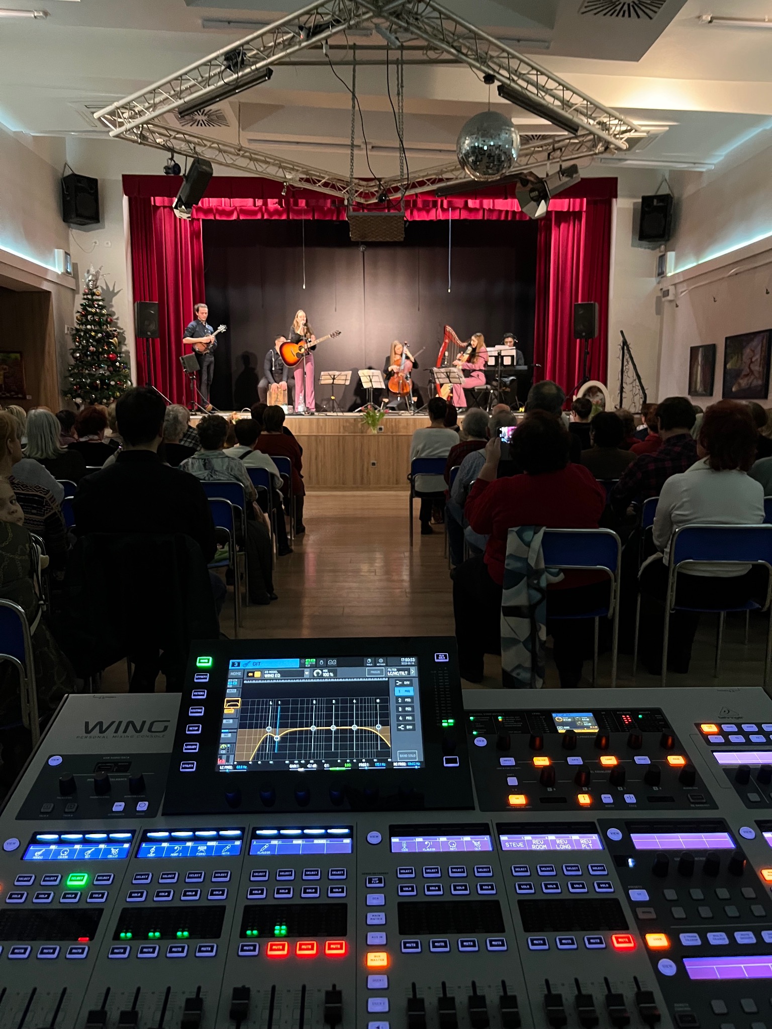 Koncert z udziałem kilku muzyków grających na instrumentach, widok zza stołu mikserskiego z widoczną publicznością i choinką w tle.