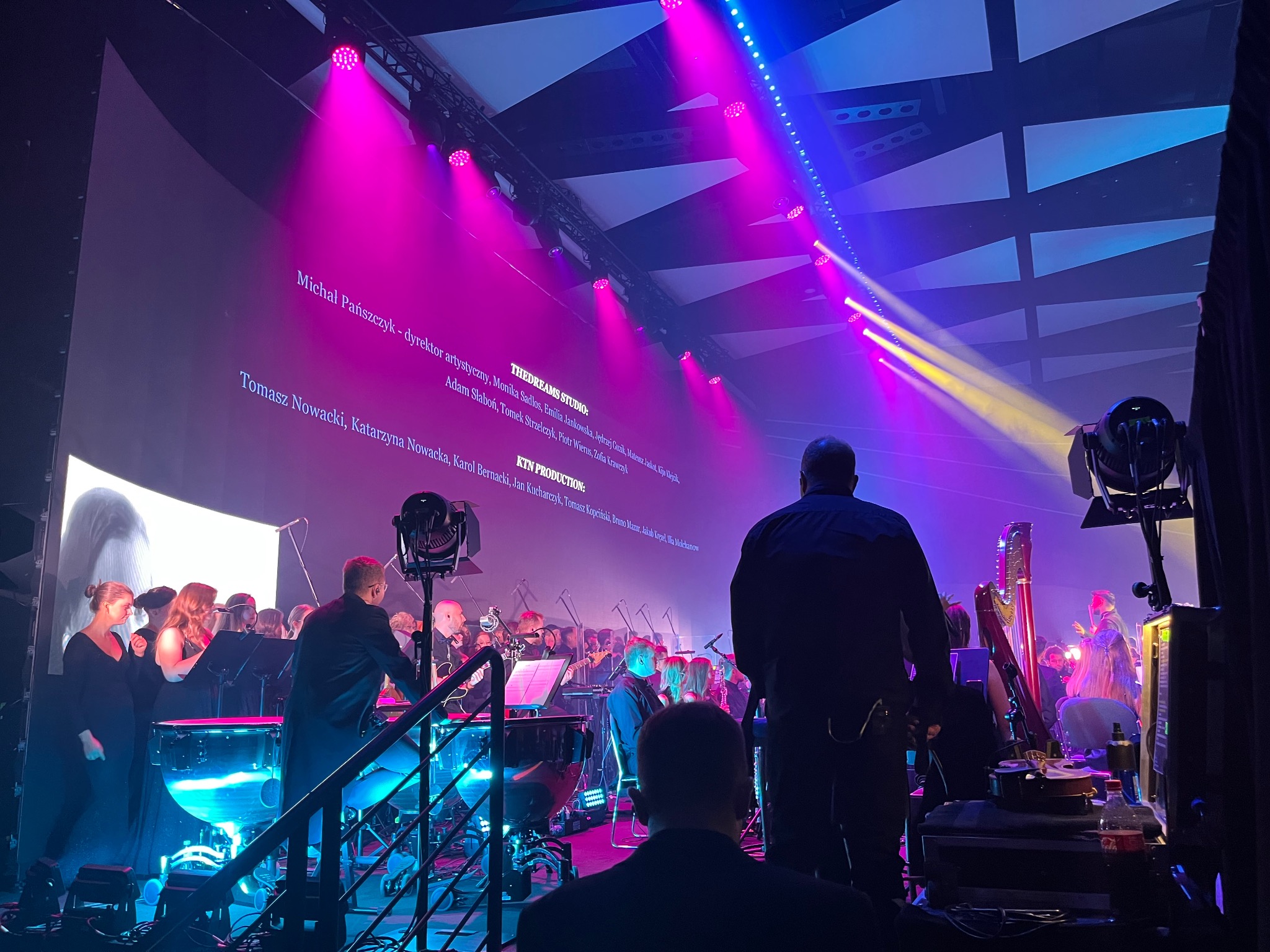 Orkiestra symfoniczna podczas występu na scenie oświetlonej kolorowymi reflektorami, widok zza kulis z widocznym osprzętem technicznym i ekranem z wyświetlanymi nazwiskami.