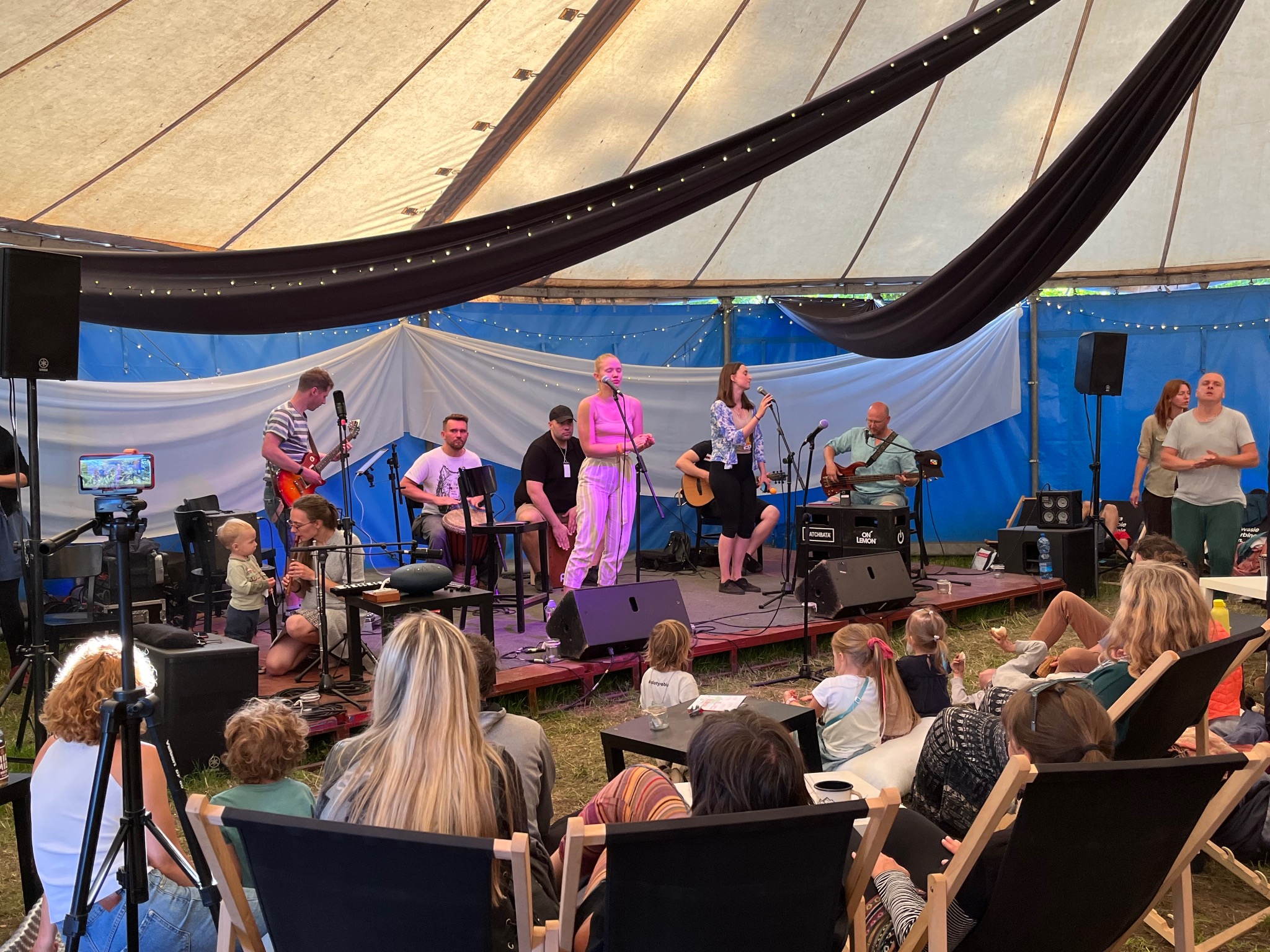 Zespół muzyczny z dwiema wokalistkami, gitarzystą, basistą i perkusistą występuje na scenie pod namiotem, przed publicznością siedzącą na leżakach.