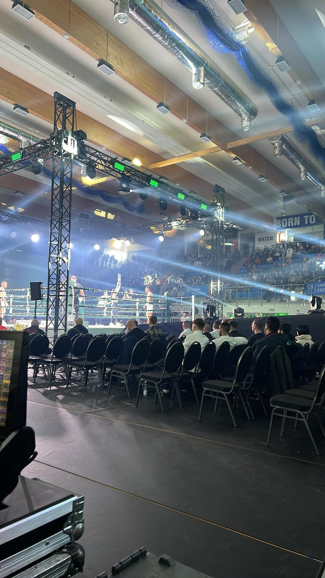 Widok zza kulis na ring bokserski podczas wydarzenia sportowego z publicznością, oświetlony reflektorami i nagłośniony, z widocznym sprzętem technicznym.