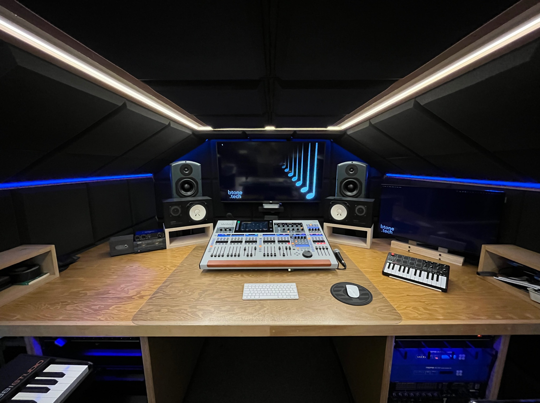 Profesjonalnie wyposażone pomieszczenie z akustycznymi panelami na ścianach i suficie, centralnie umieszczony mikser audio z licznymi suwakami i pokrętłami, monitory studyjne na drewnianych...