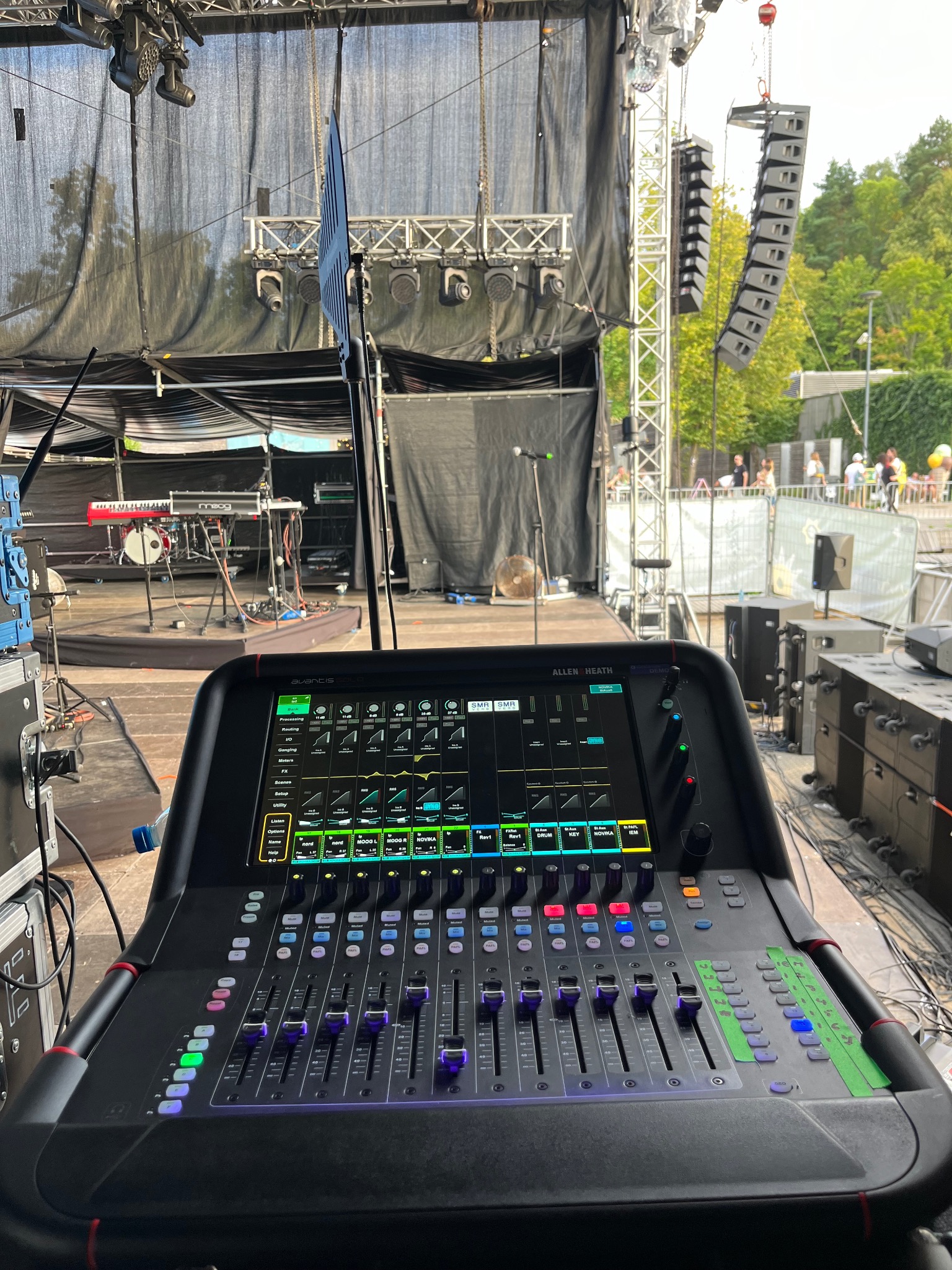 Konsoleta mikserska Allen & Heath Avantis Solo na tle sceny plenerowej z nagłośnieniem liniowym i instrumentami muzycznymi, widok zza urządzenia.